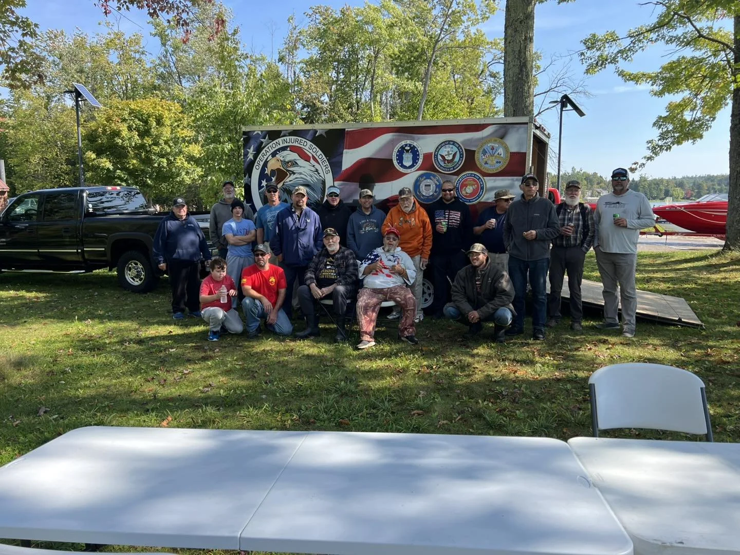 Group of people posing outdoors in front of a truck with military and patriotic logos and an eagle. Trees and a red speedboat are visible in the background.