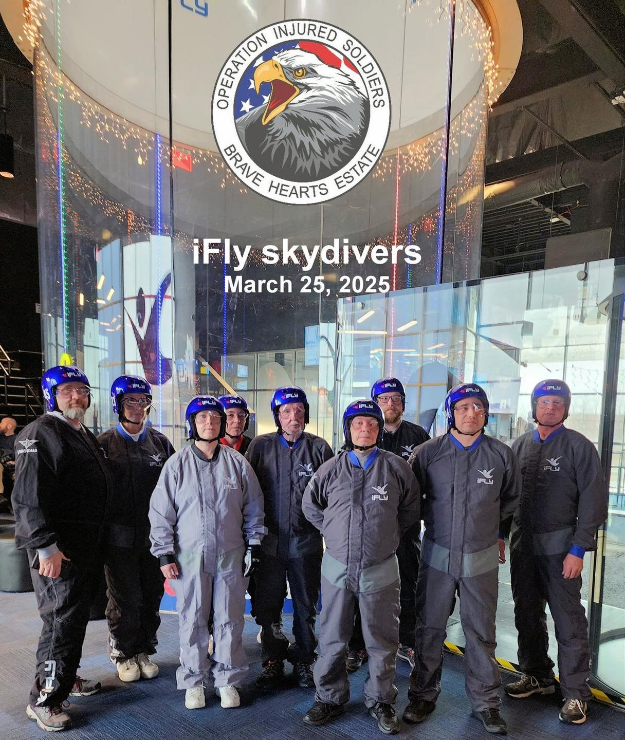 Group of skydivers in jumpsuits and helmets standing in front of a glass wall at an indoor skydiving facility celebrating a skydiving event on March 25, 2025.