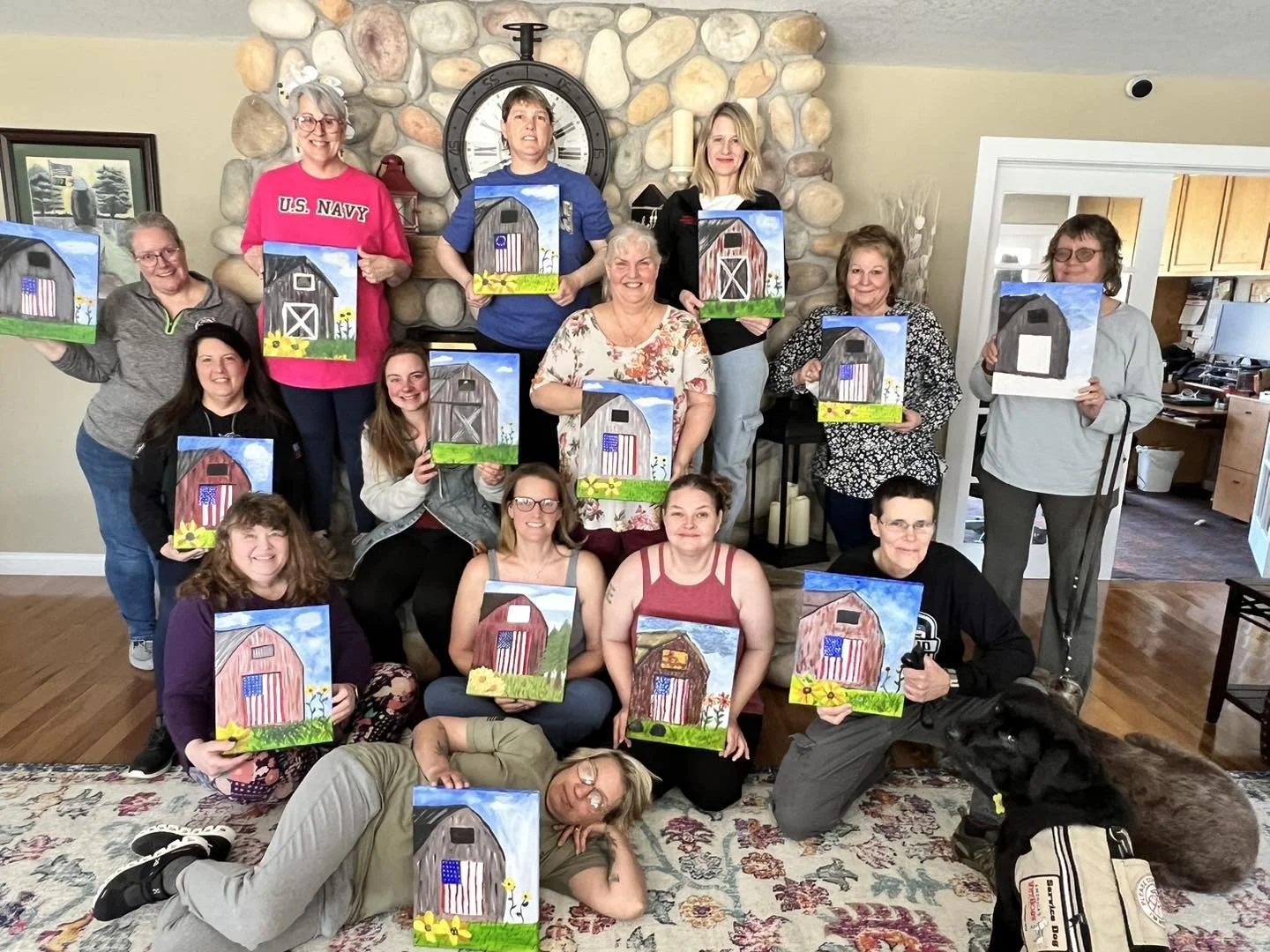 A group of people in a living room holding paintings of barn scenes, with some featuring American flags. They are smiling and posing for a photo, with a stone fireplace and clock in the background.