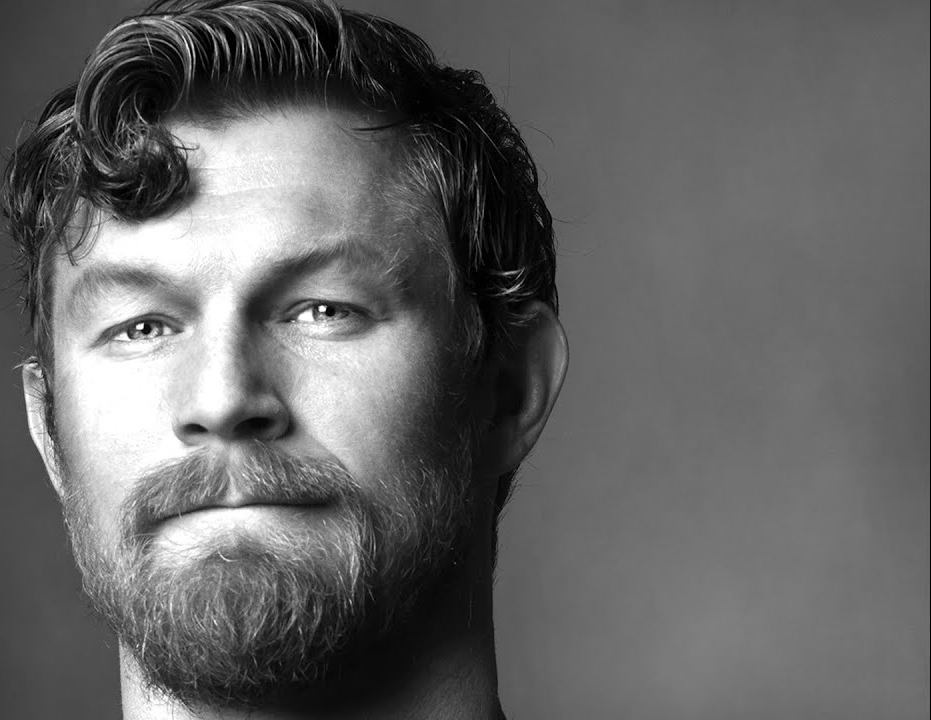 Black and white close-up portrait of a man with wavy hair, beard, and mustache, looking directly at the camera with a slight smile.