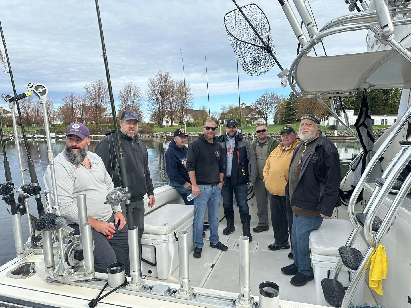 Group of eight men standing on a boat by a calm water body with houses and trees in the background, some holding fishing rods.