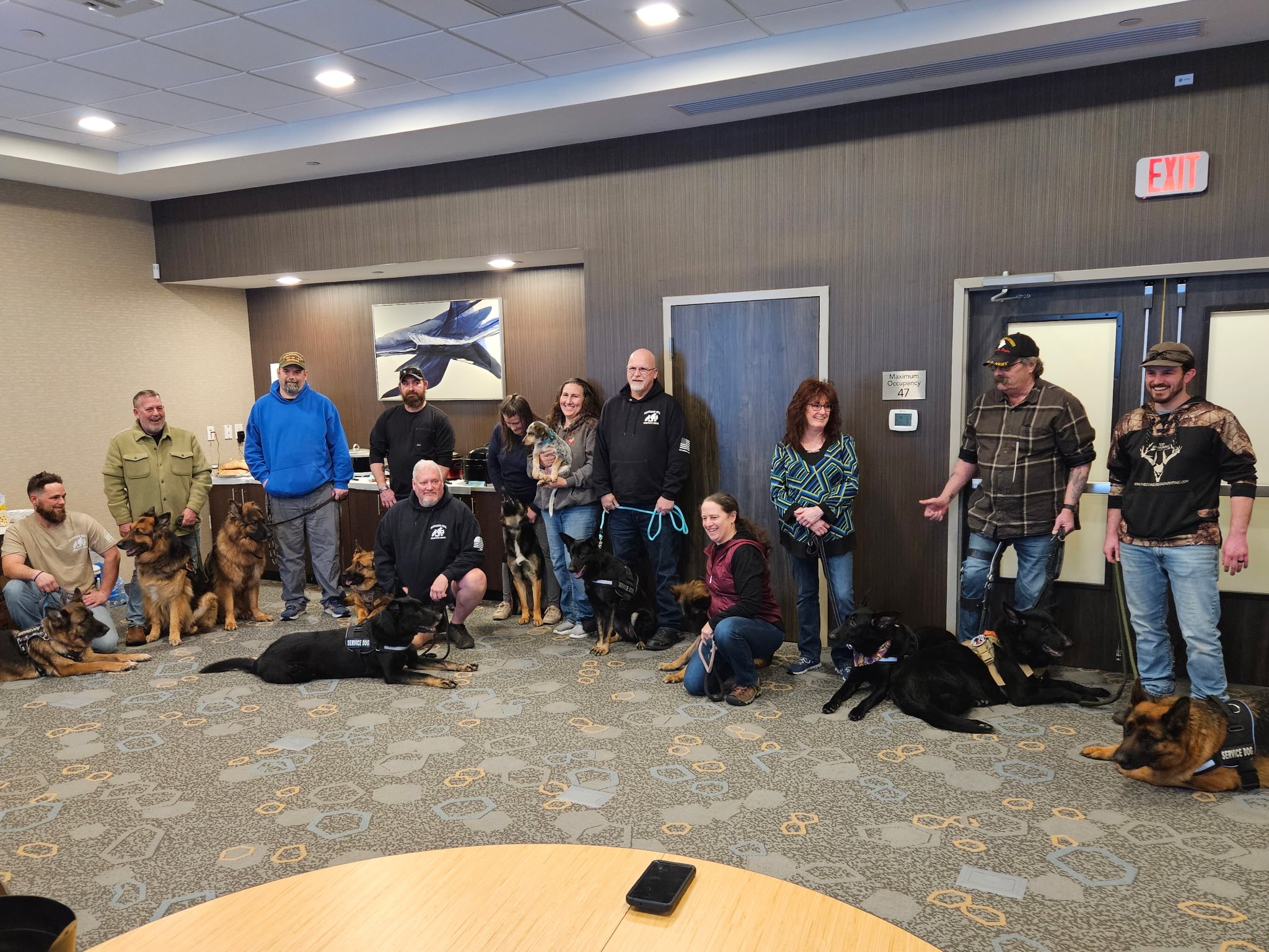 A group of people and dogs gathered in a room, some sitting and some standing, with a brown wall, artwork, and an exit sign in the background.