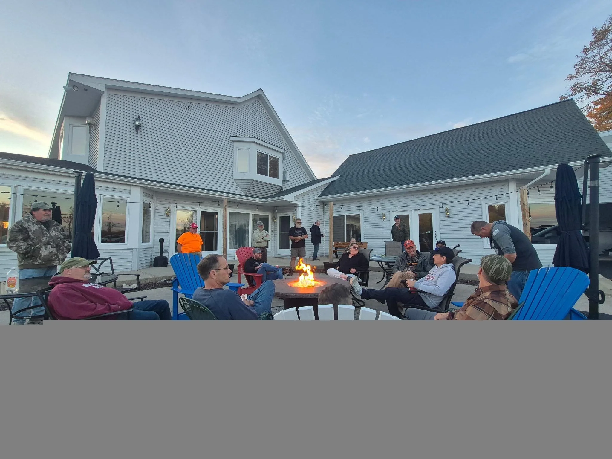 People gathered outdoors on a patio around a fire pit, socializing, with a white house and clear sky in the background.