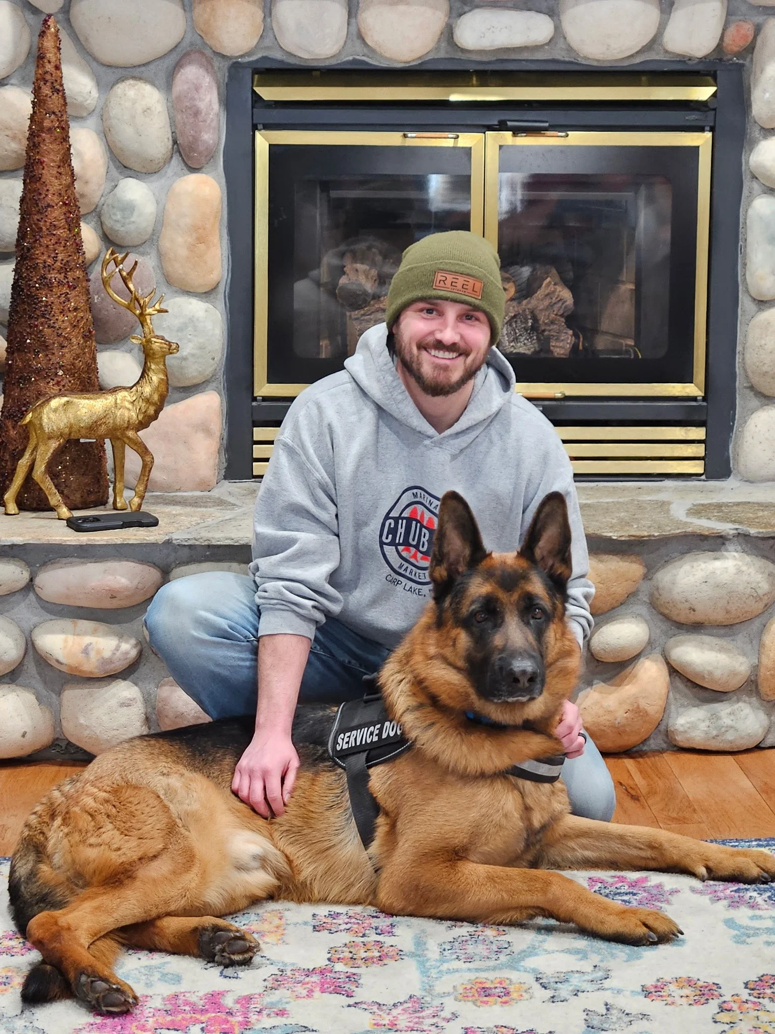 A man with a beard and a green beanie sitting on the floor with two German Shepherd dogs in front of a stone fireplace. The man is smiling, wearing a gray hoodie, and has a service dog vest on one of the dogs. The fireplace has a glass door, and ther