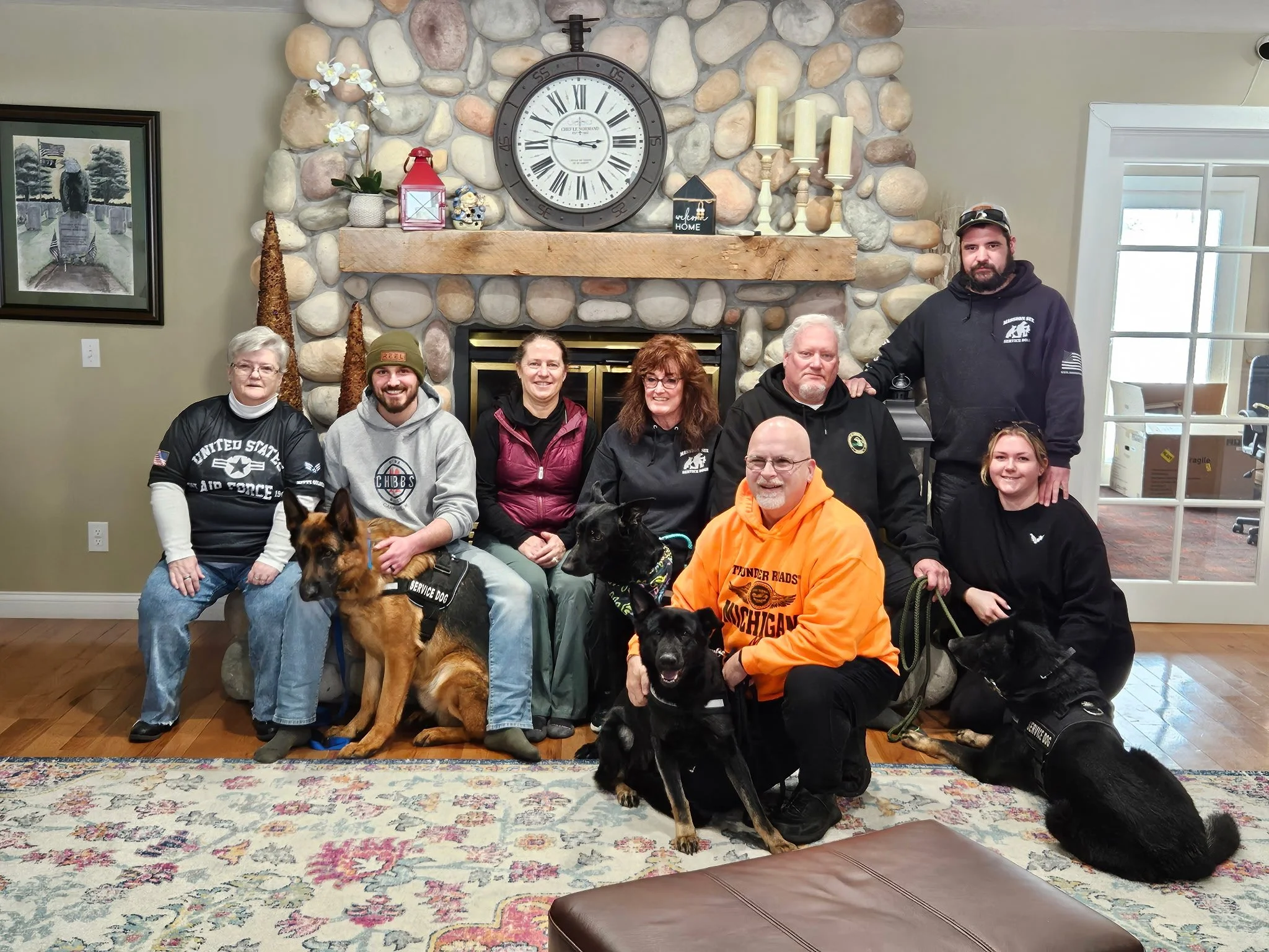 A group of eight people and four dogs posing in a living room with a stone fireplace and large clock above it. People are sitting and standing, smiling at the camera. The room has a patterned rug, decorative candles, and a plant on the mantle.