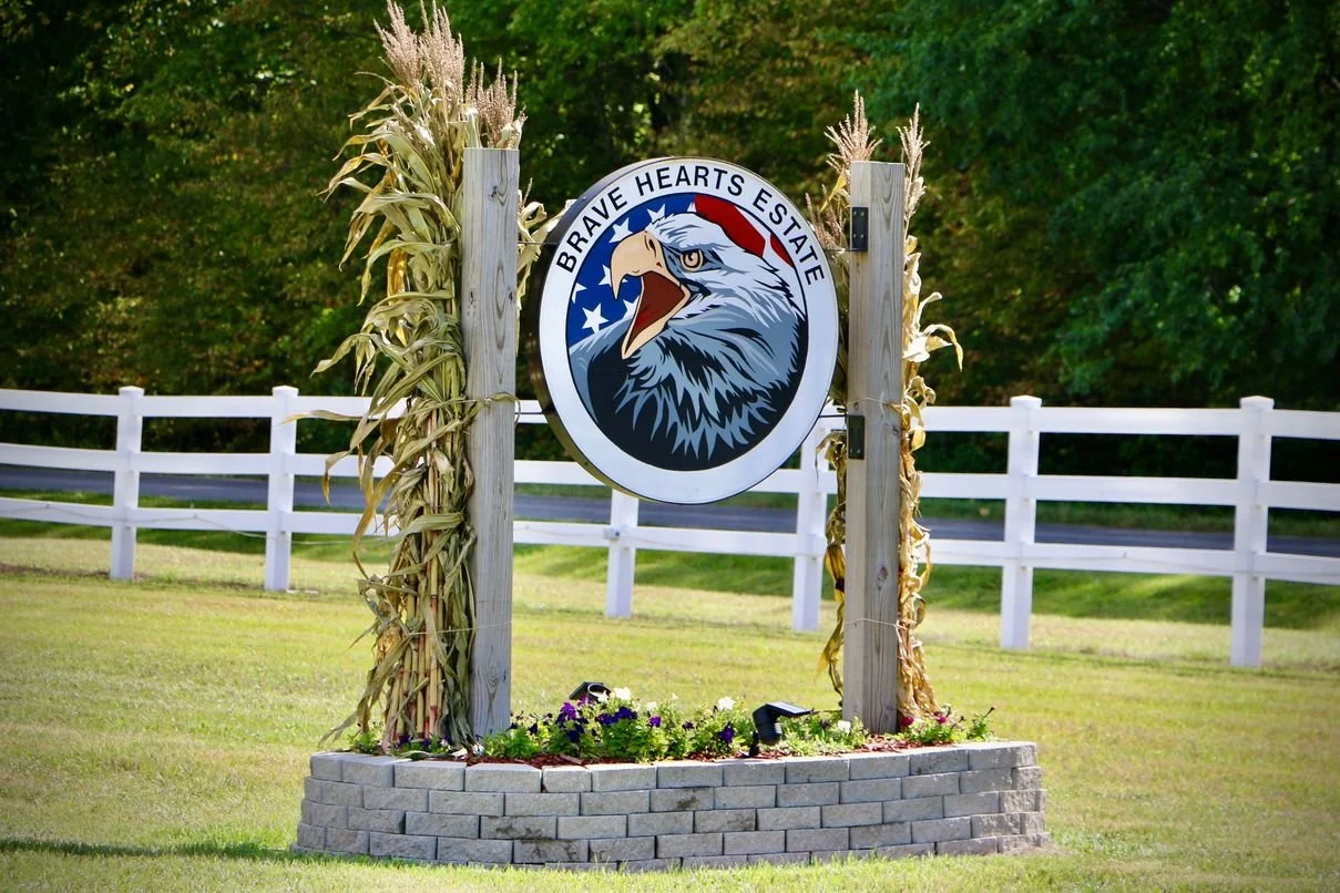 A sign for Brave Hearts Estate featuring an eagle with an American flag-themed background, surrounded by dried cornstalks and a small flower garden, with a white fence and green trees in the background.