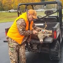 A person dressed in camouflage clothing and an orange safety vest holding a deer by its head next to a pickup truck in a rural outdoor setting.