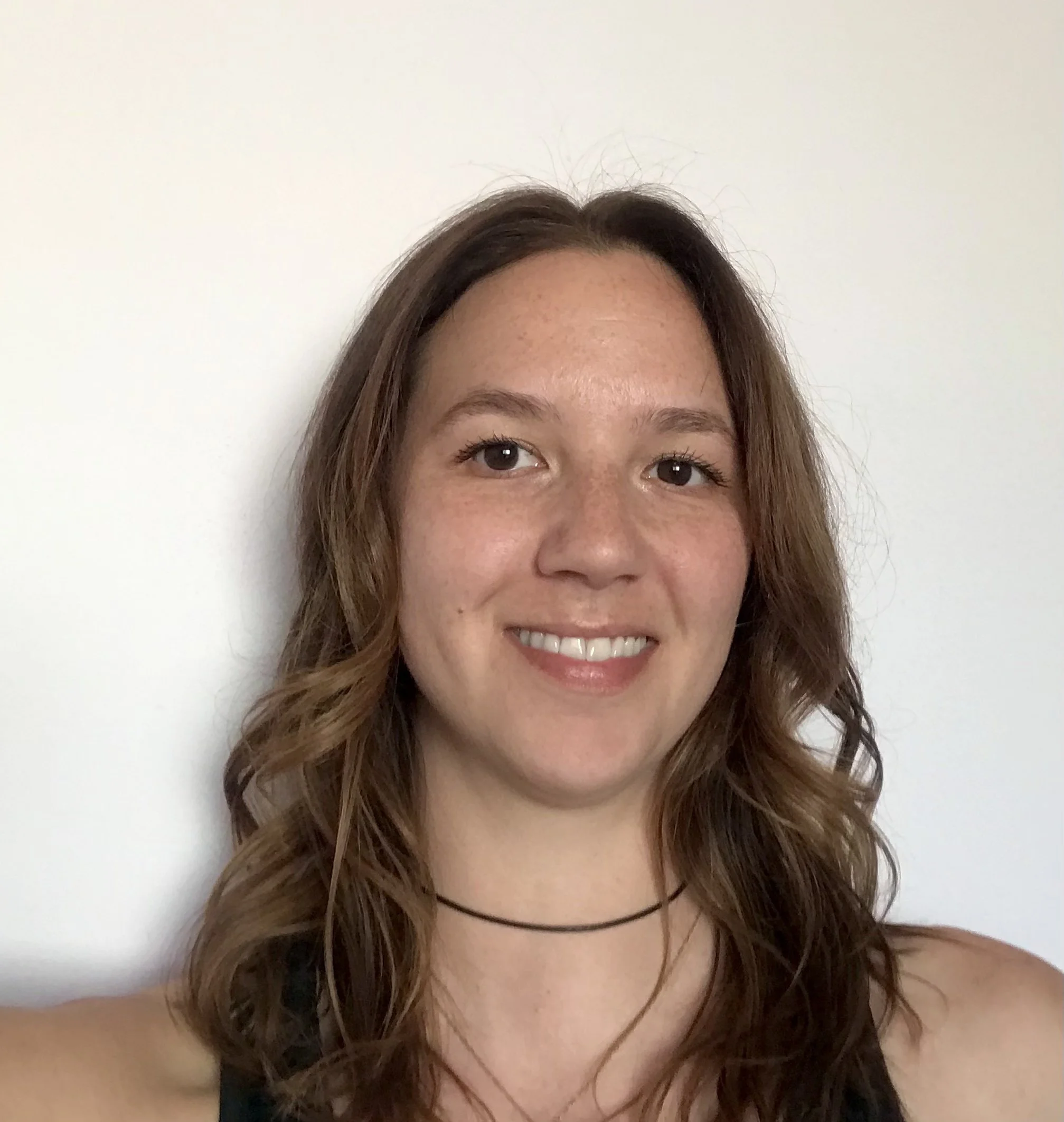 A young woman with wavy brown hair, smiling in front of a plain white wall.