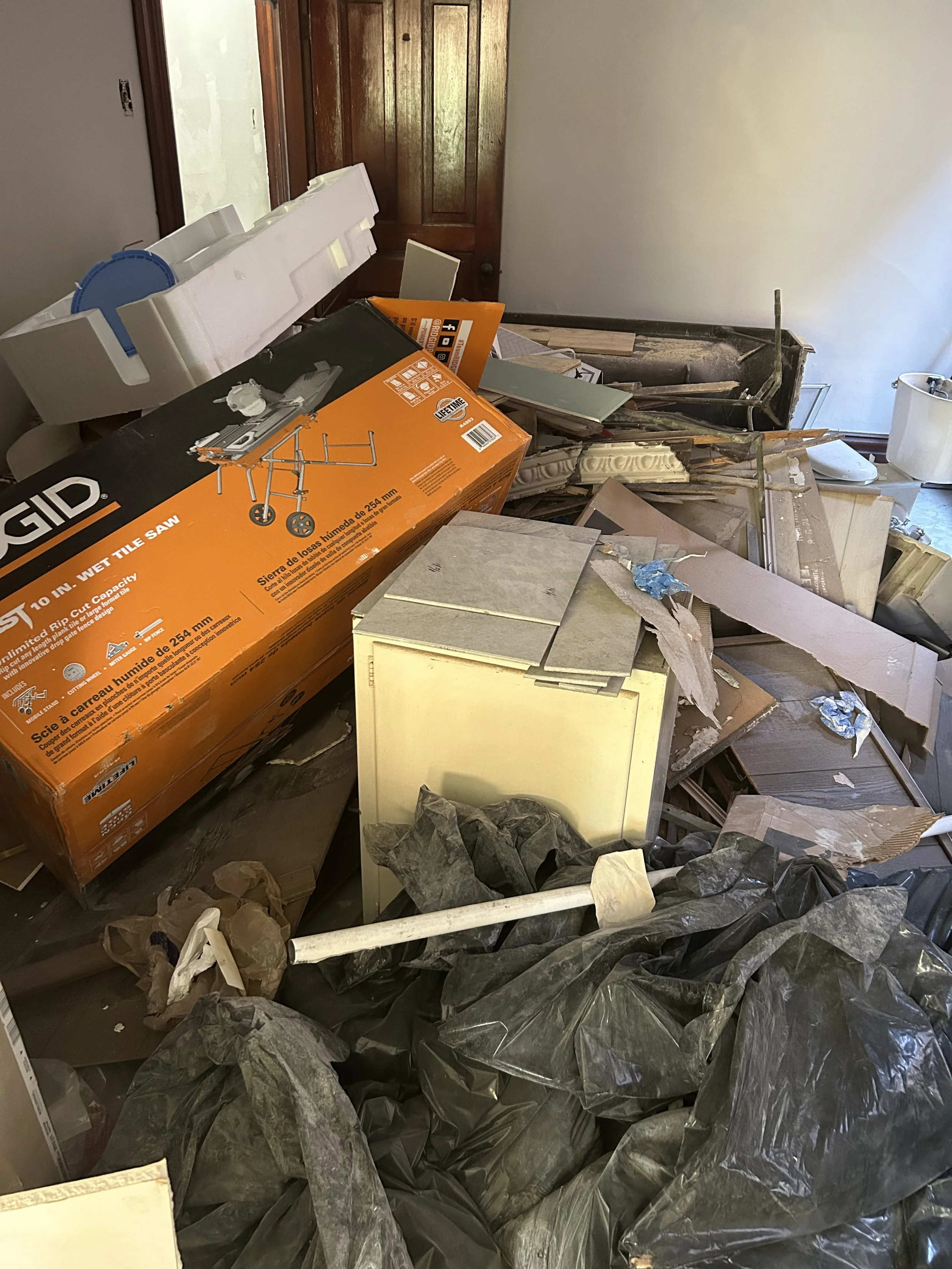 A room filled with construction debris, including black trash bags, a box labeled 'Wet Tile Saw,' pieces of drywall, and various construction materials, with a wooden door in the background.