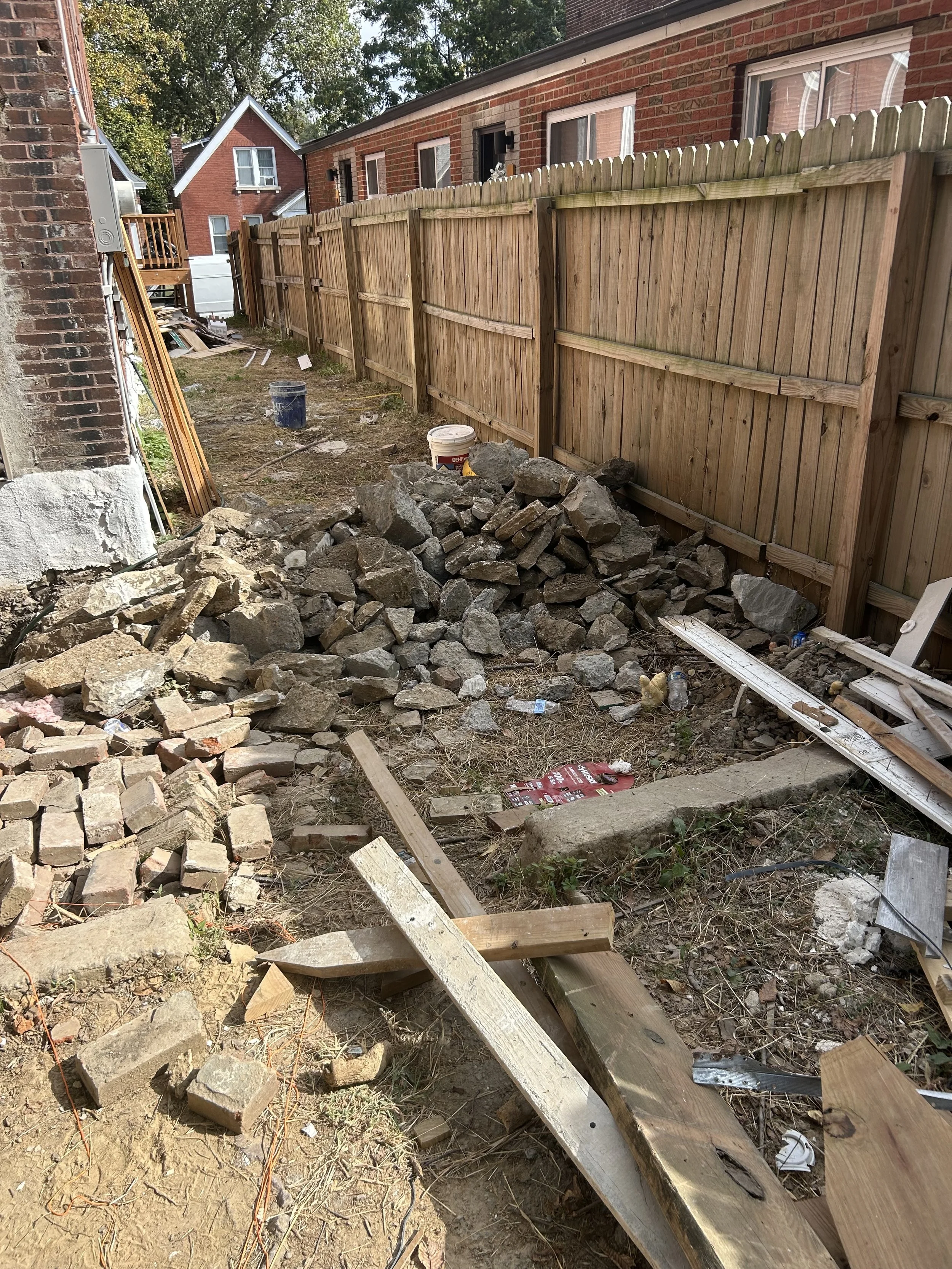 Backyard with a wooden fence, construction debris, rocks, bricks, wood planks, pipes, and building materials, indicating ongoing yard work or renovation.
