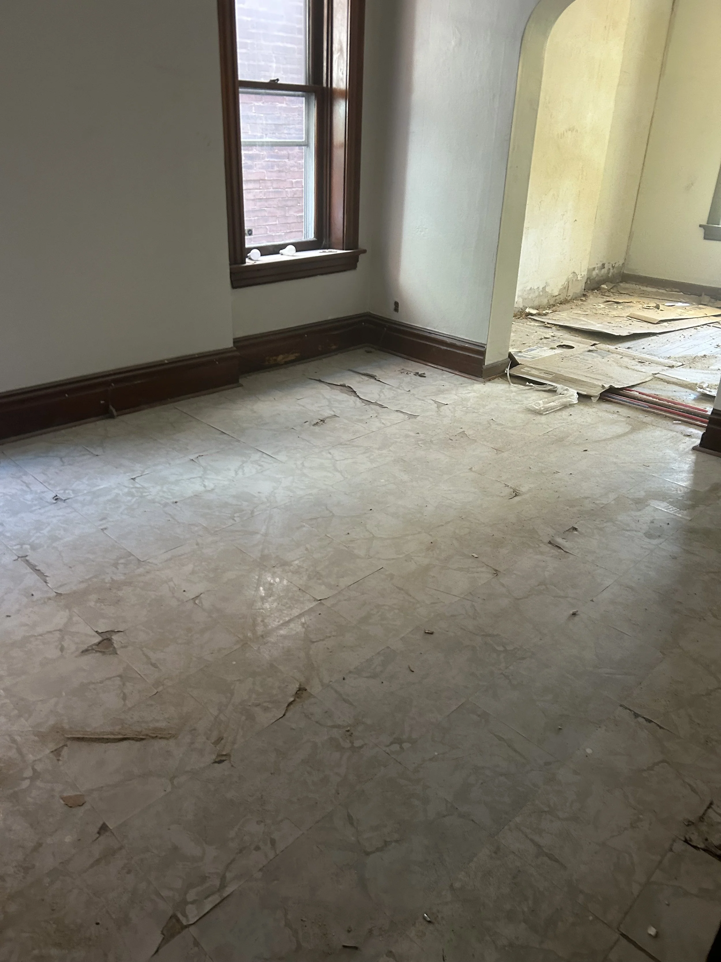 Room with damaged, peeling tile floor, window with wooden trim, and wall with signs of water damage or mold.