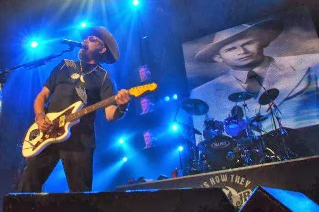 Musician playing guitar on stage with blue lights, wearing sunglasses and a hat, large black and white photo of a man in a hat projected on screen behind him, drum set visible.