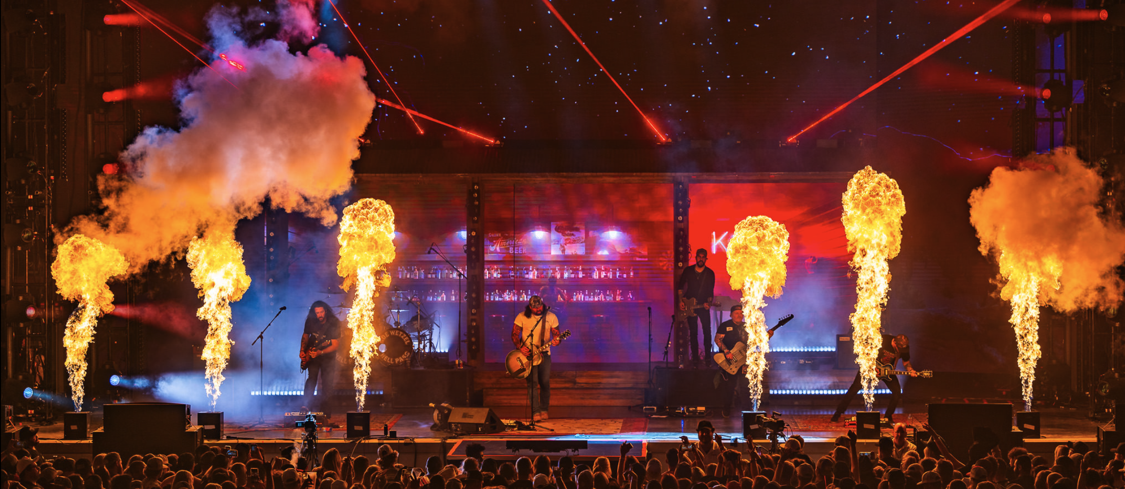 A live concert with flames and smoke effects on stage. Musicians are performing with a crowd watching.