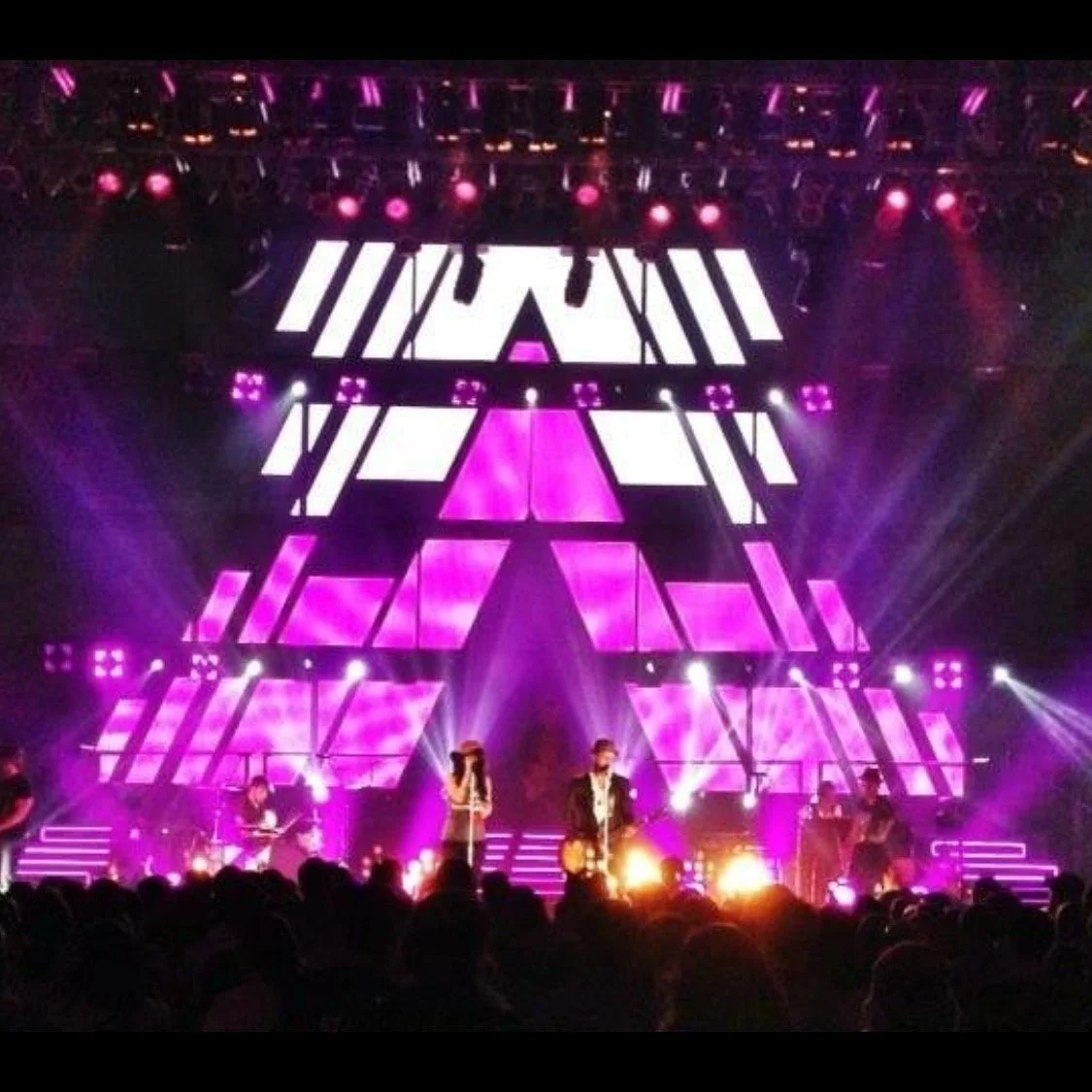 Music concert stage with vibrant pink and white lighting, performers including a singer and guitarist, and a crowd in the foreground.