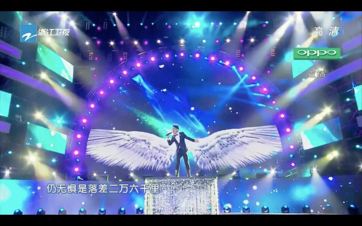 A male singer in a suit stands on a sparkling platform with large angel wings projected on the LED background, performing on a brightly lit stage with colorful lights and screens