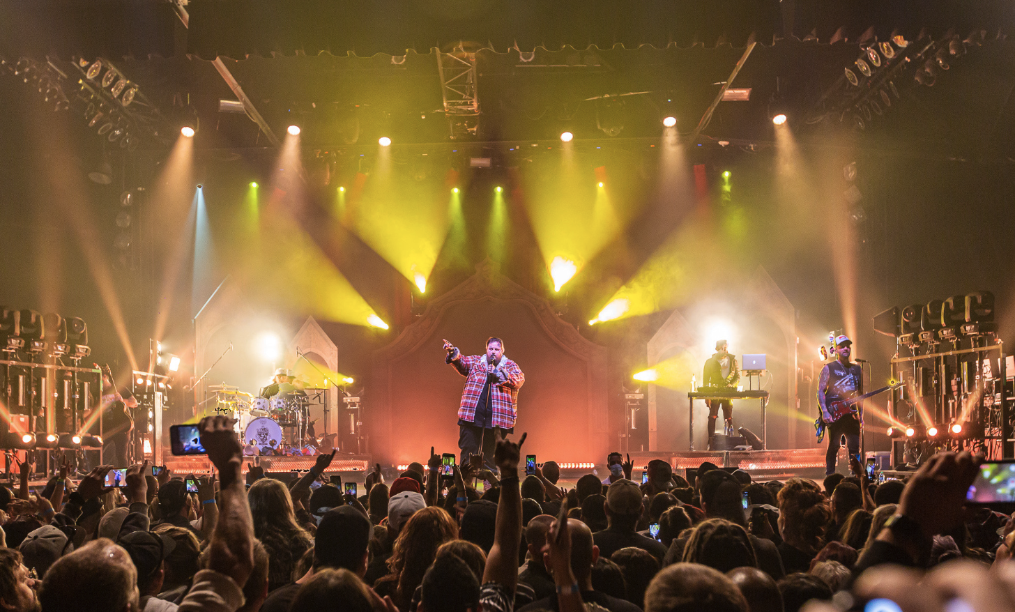 A band performs on stage during a concert with bright yellow and white lights, an audience taking photos, and a lead singer in a plaid shirt at the center.