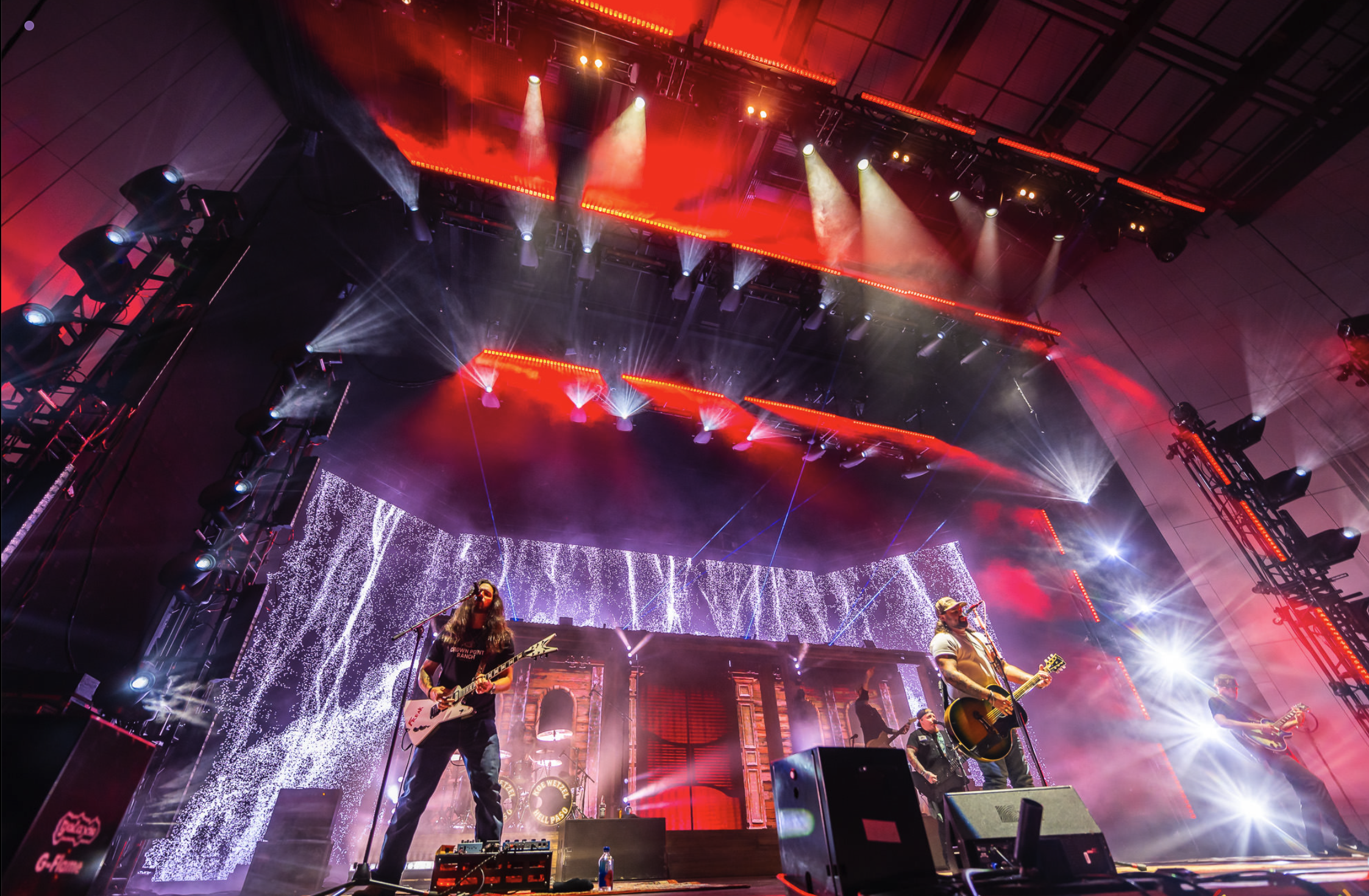 A live music concert on a stage with bright red and white lights, musicians playing guitars, and a large LED screen showing visual effects in the background.