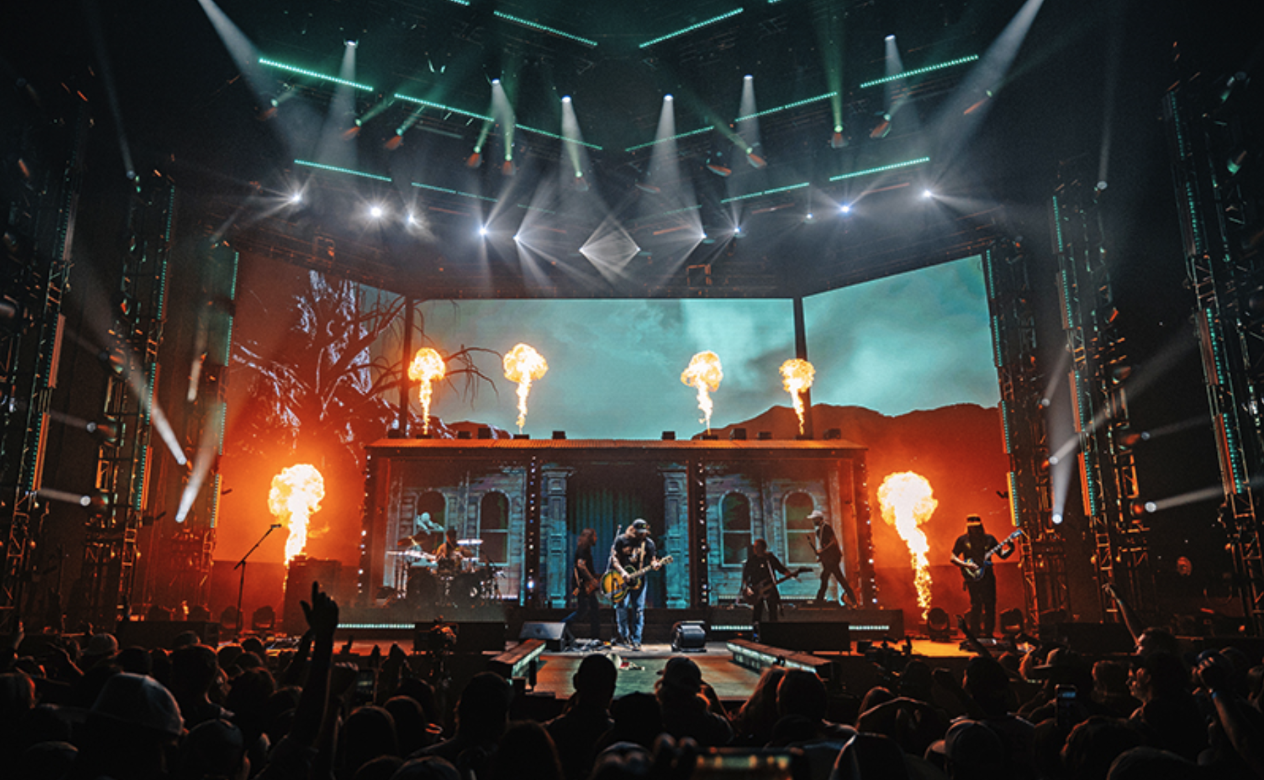 A live concert with a band playing on stage, pyrotechnics shooting flames, and an animated backdrop with trees and a sky, surrounded by vibrant stage lighting.