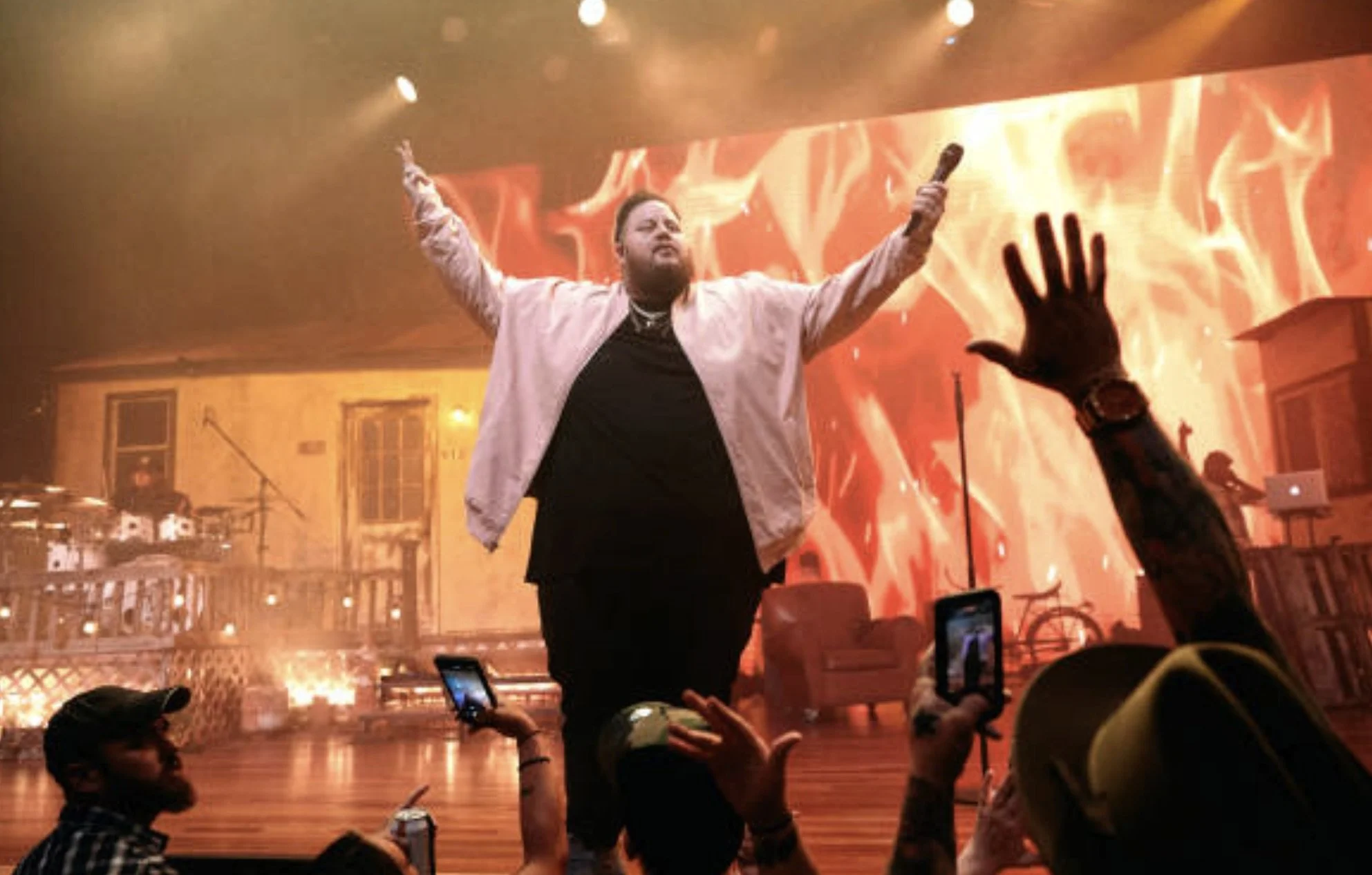 Performer with arms raised on stage during concert, audience reaching out, in a venue with warm lighting and a fiery background on a large screen.
