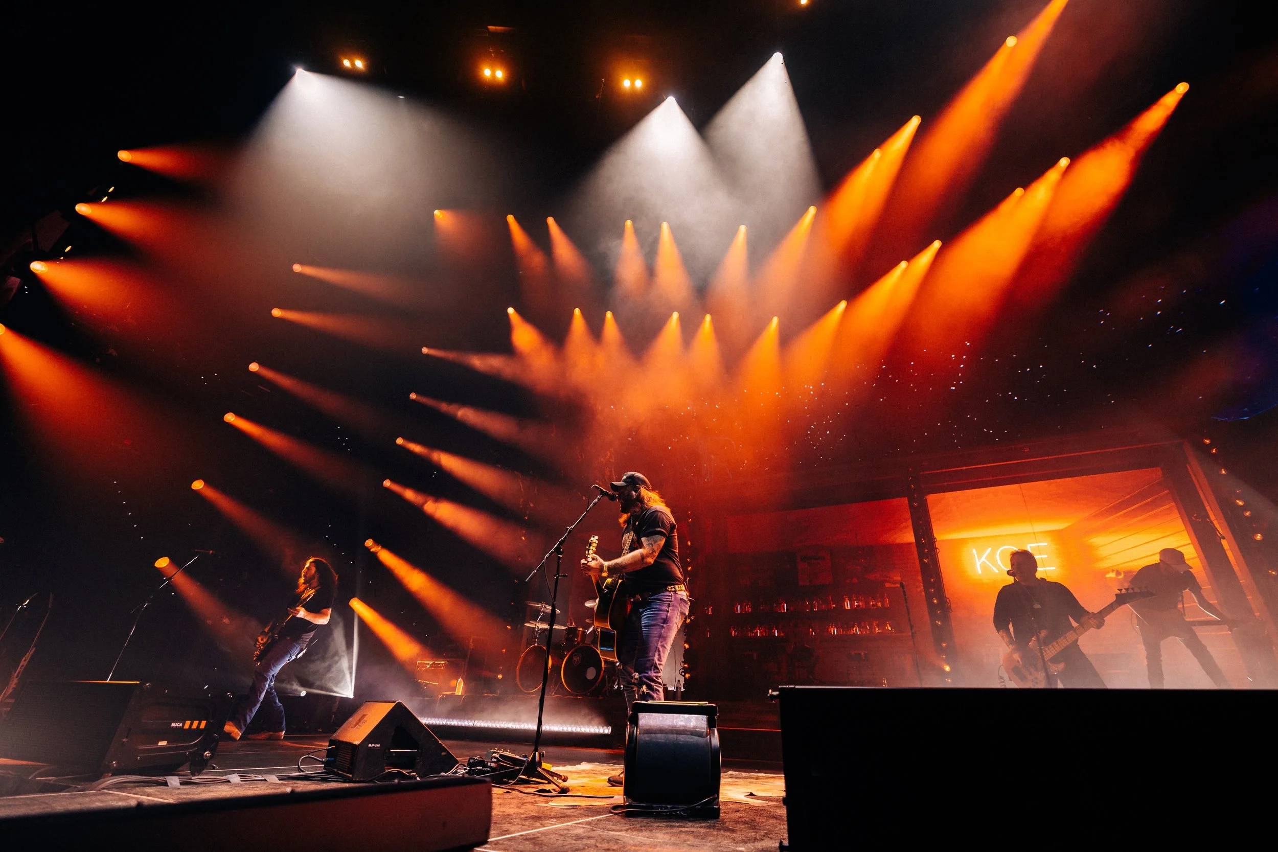 A live concert scene featuring musicians on stage surrounded by orange and white stage lights. One musician is playing an electric guitar and singing into a microphone. The background displays a bar and fans in silhouette at the front of the stage.