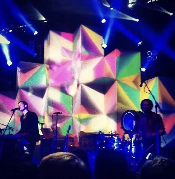 Musicians performing on stage with a colorful, geometric light display in the background.