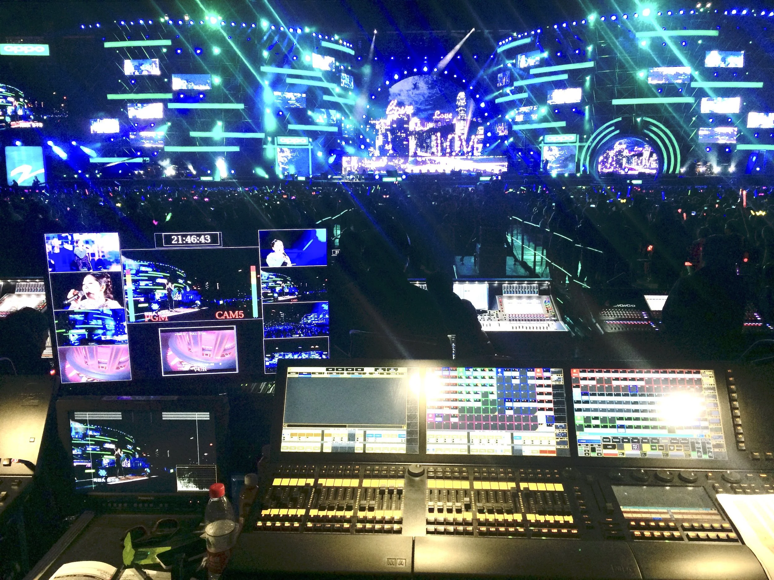 A large stage at a concert or event with vibrant green, blue, and white lighting effects, multiple screens displaying visuals, and an audience with glow sticks. The digital control consoles and equipment are visible in the foreground.