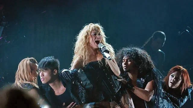 A woman with curly blonde hair singing into a microphone on stage, surrounded by three women with dark curly and straight hair and a woman with red hair, all in dark clothing.