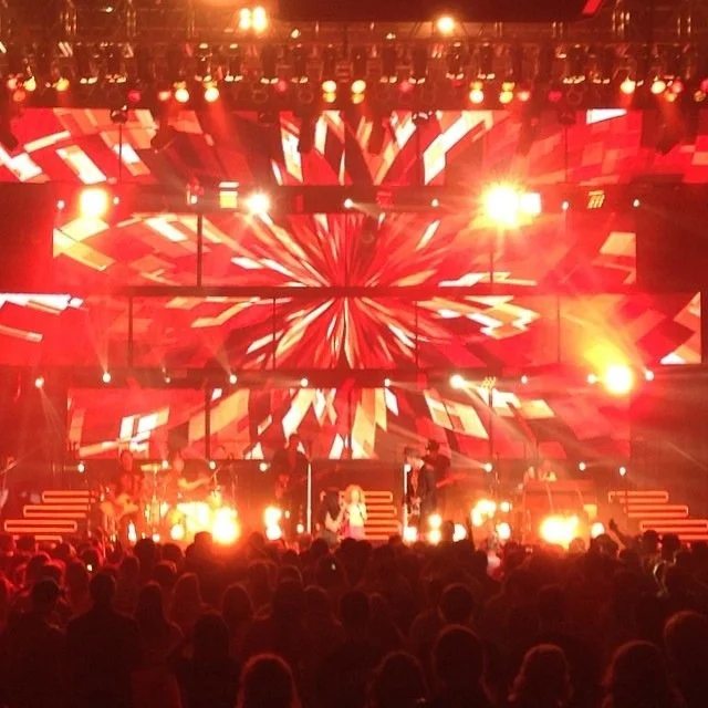 A live concert with a band performing on stage, illuminated by colorful lights and visual effects projected on a large screen behind them, with an energetic audience in front.