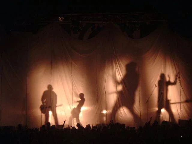 Silhouettes of four musicians performing on stage with a curtain backdrop, backlit to create shadows, during a concert.