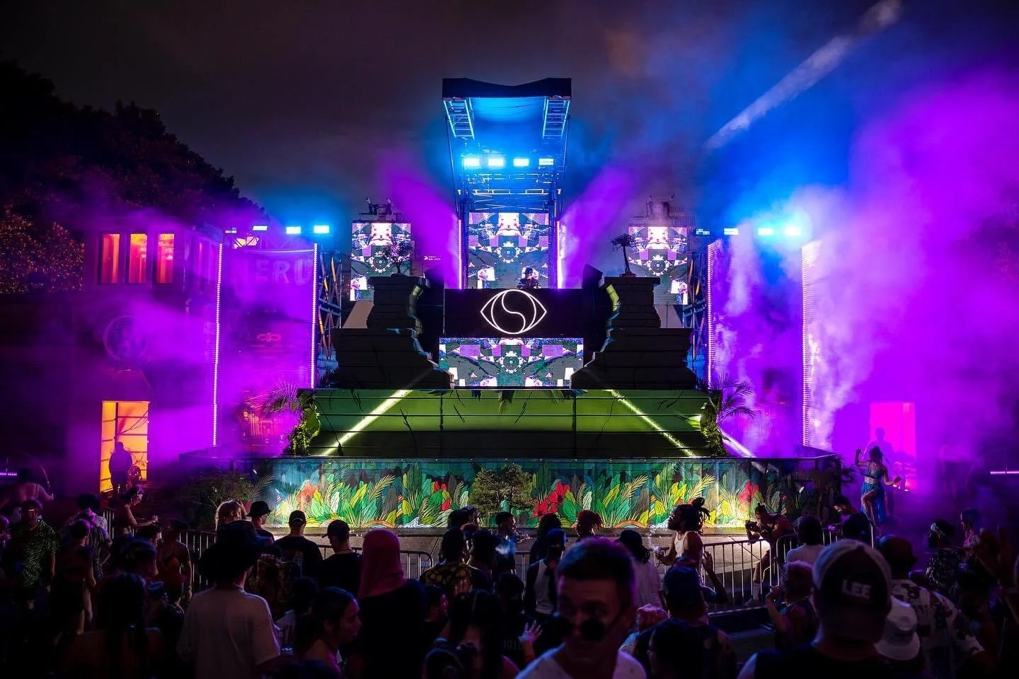 A vibrant outdoor music festival stage at night with colorful lights, smoke effects, and a crowd of attendees in front.