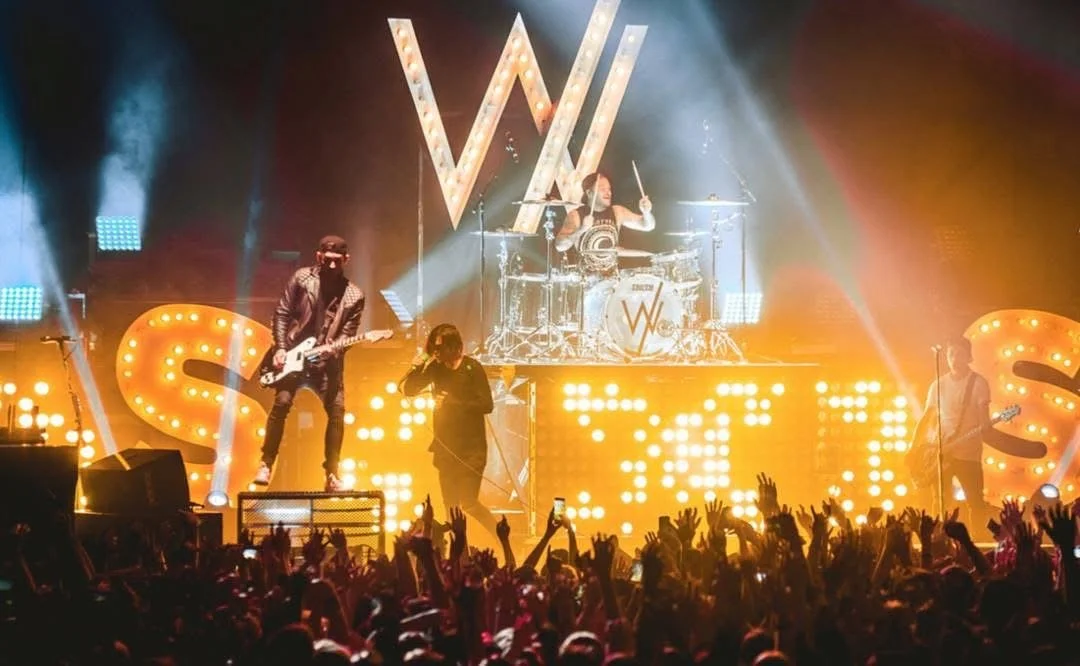 A live rock band performing on stage with large illuminated letters spelling 'WAS' in the background. The stage is lit with bright lights and smoke, and the audience has hands raised.