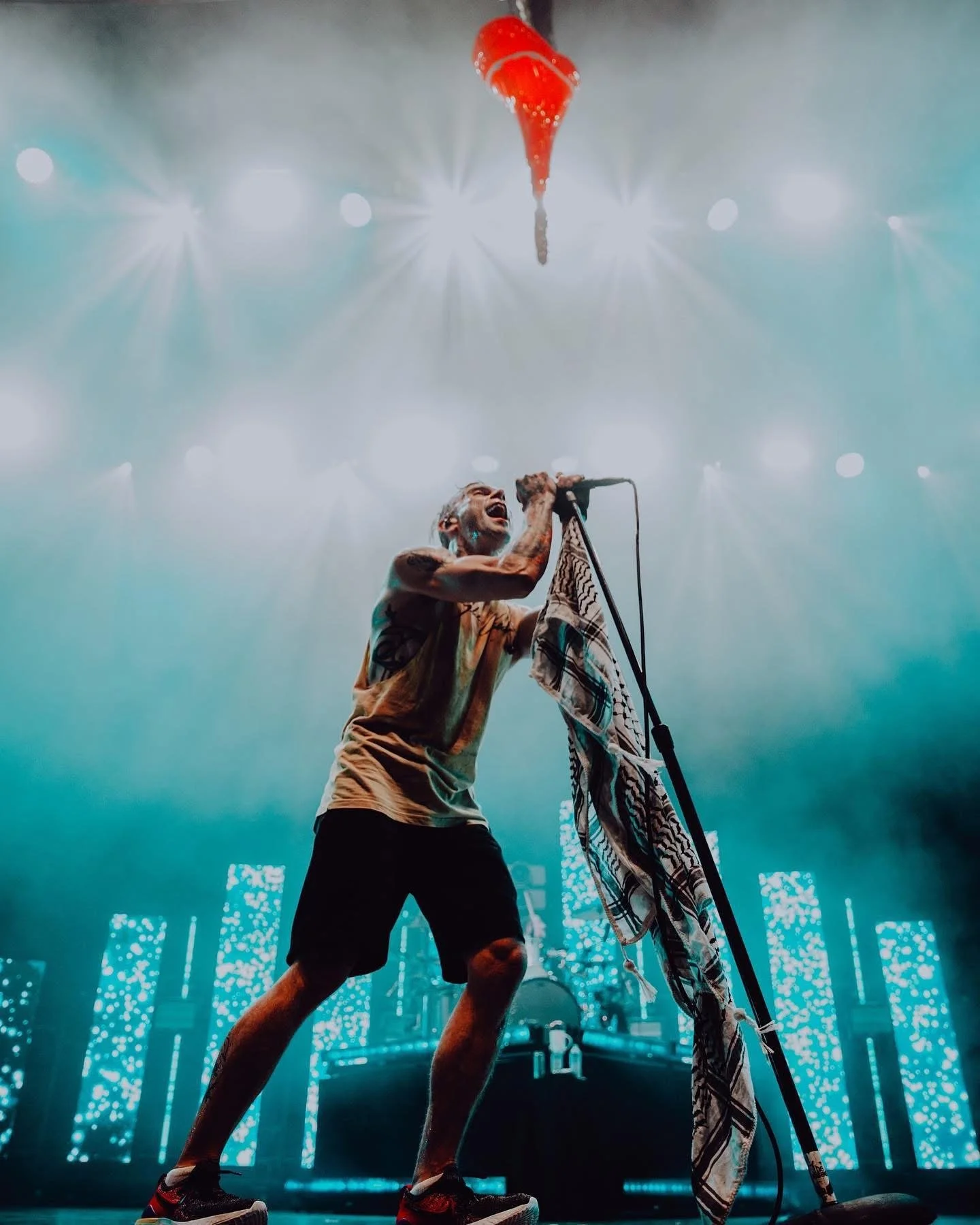 A performer singing into a microphone on stage with bright lights and LED screens in the background, holding a cloth, wearing a sleeveless shirt and shorts, with a tambourine attached to the mic stand.