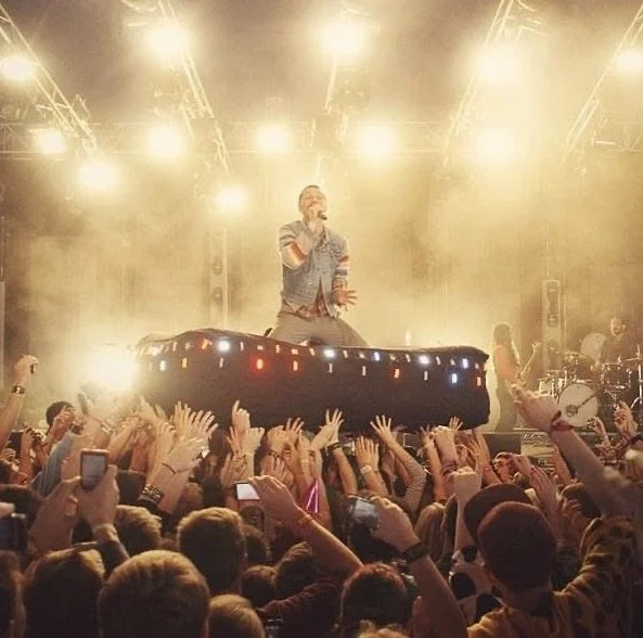 Performer on stage with DJ booth, bright stage lights, and a crowd cheering and taking photos at a concert.