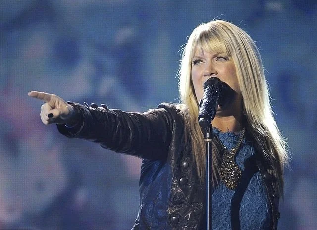 A woman with blonde hair singing into a microphone on stage, pointing with her right hand.