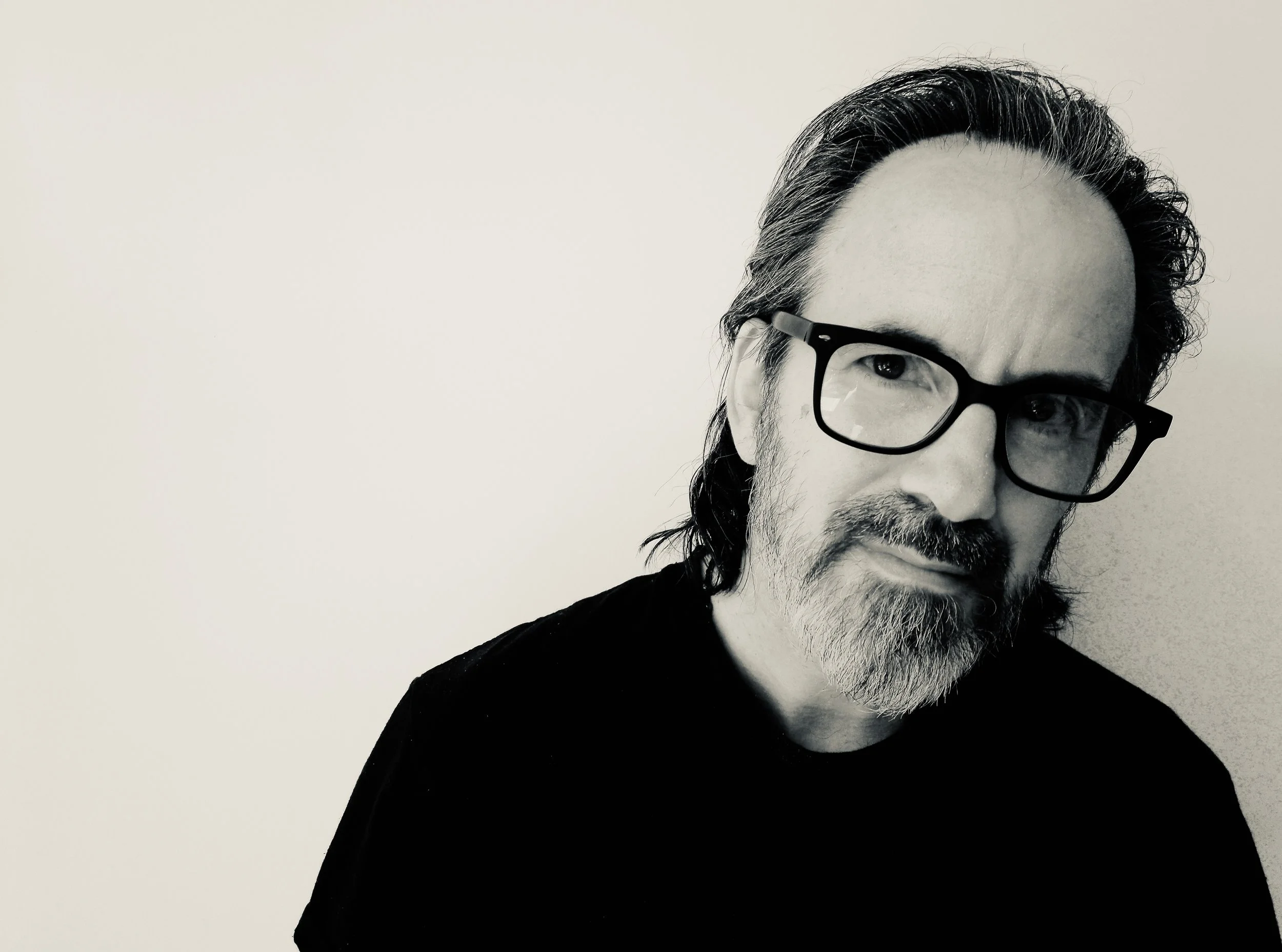 A black and white portrait of a man with glasses, long wavy hair, a beard, and mustache, wearing a dark shirt, against a plain light background.
