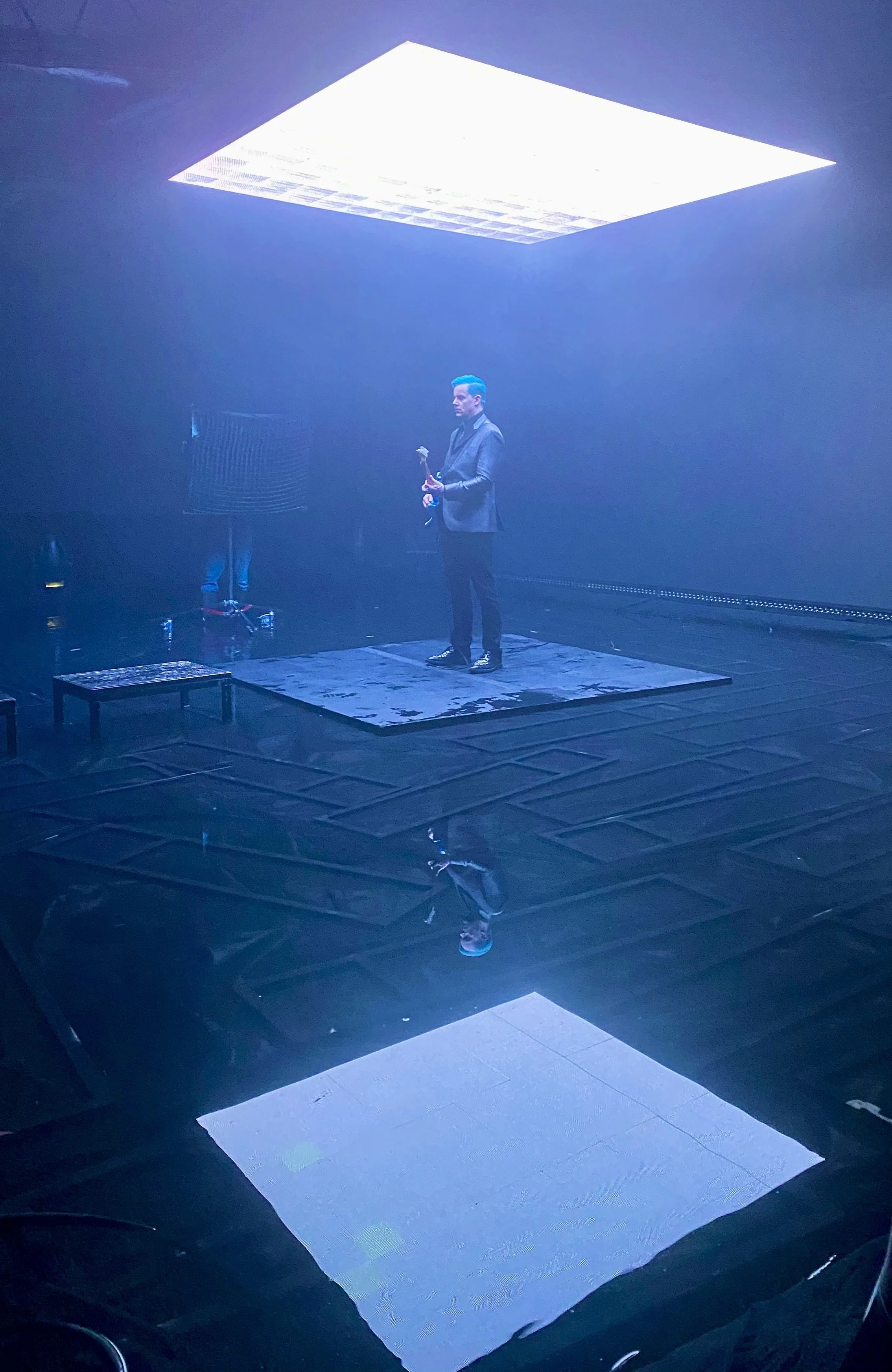 A man in a gray suit holding a microphone stands on a platform made of cardboard or foam boards. There is a large illuminated rectangular ceiling panel above him, casting bright white light. The environment is dark and foggy, possibly an indoor event or performance space.