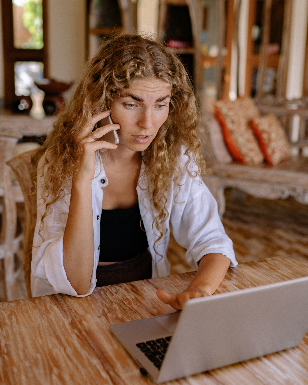 A young woman with curly blonde hair talking on a cell phone while sitting at a wooden table and looking at a laptop.