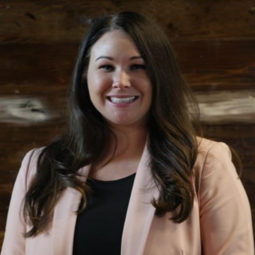 A woman with long, wavy dark hair smiling, wearing a light-colored blazer over a black top, standing against a wooden background.