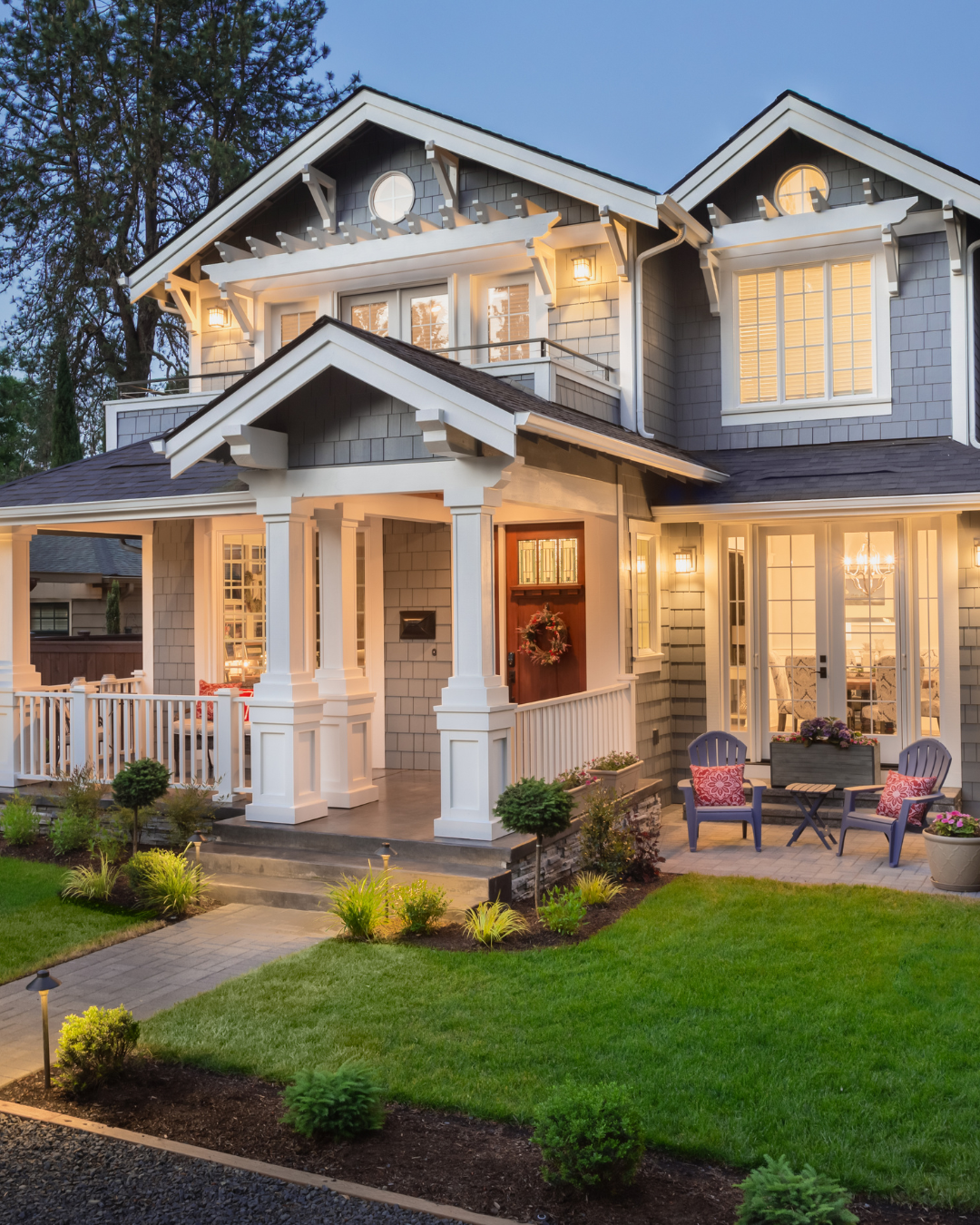 A two-story house with a porch and outdoor seating, lit up during evening with a well-kept lawn and garden.
