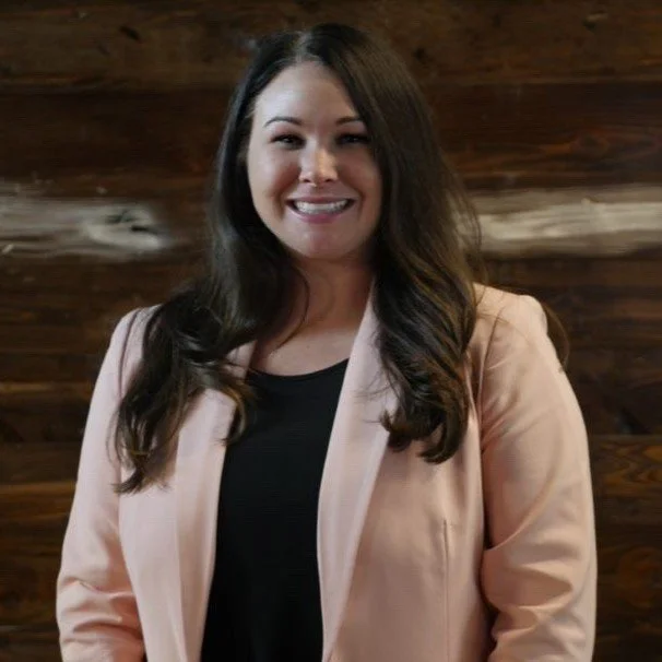Smiling woman with long dark hair, wearing a light-colored blazer over a black top, standing in front of a wooden wall.