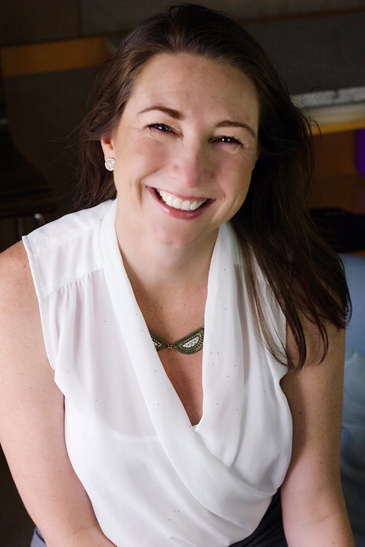 A smiling woman with brown hair, wearing a white sleeveless top and a decorative necklace.