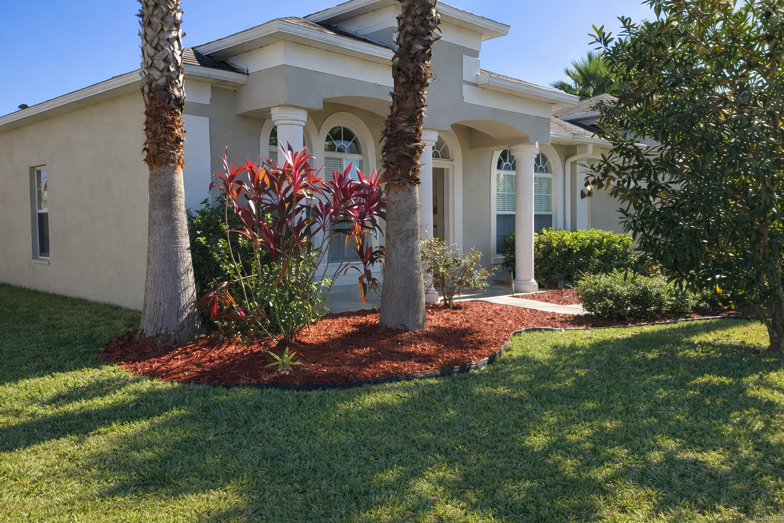 Front yard of a house with two palm trees, red mulch, green shrubs, and a manicured lawn