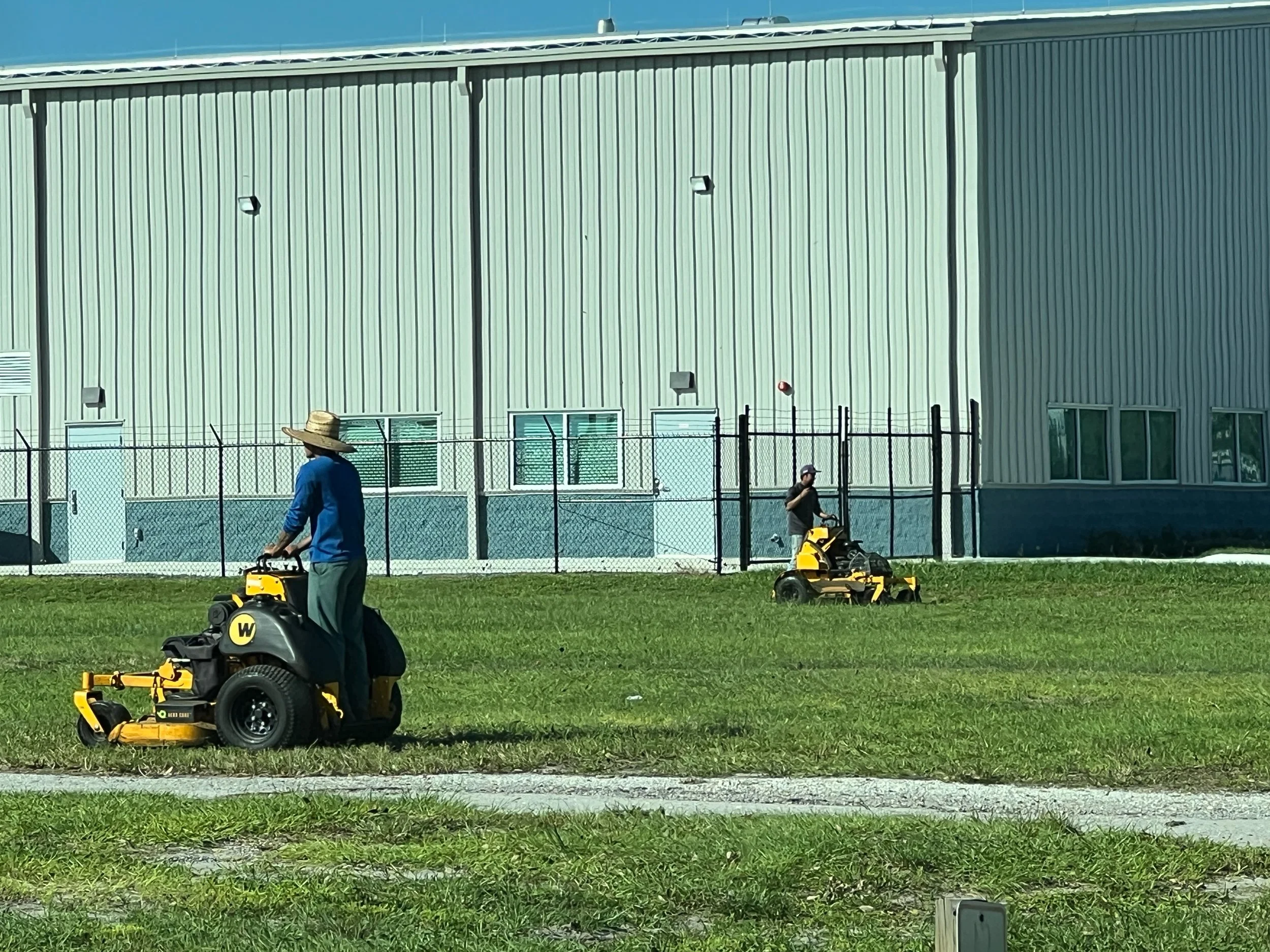 Two men mowing grass with riding lawn mowers in front of a large industrial building, with one man in a blue shirt and straw hat, and the other in a dark shirt and cap.