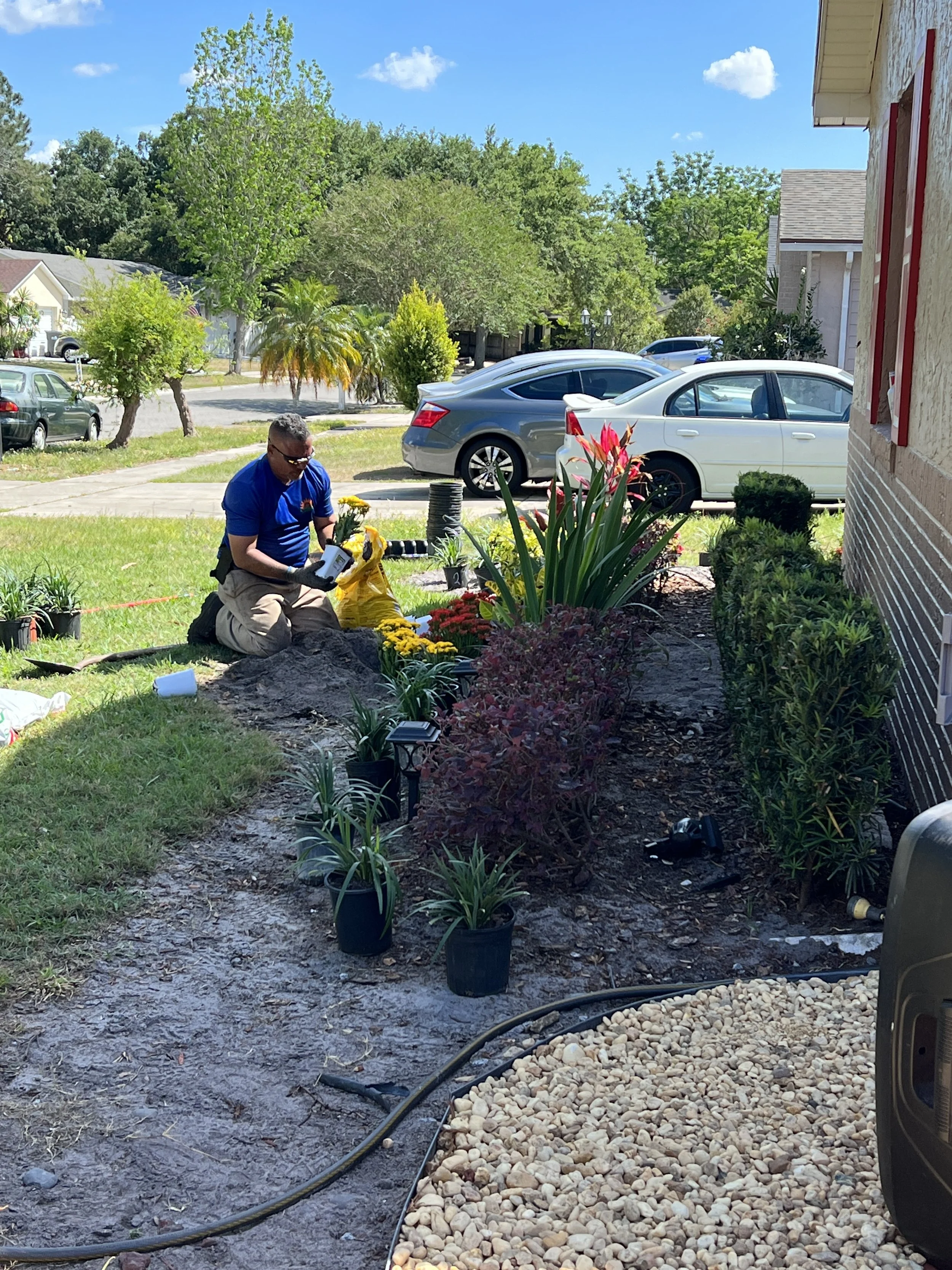 A man kneels on the ground planting flowers in a garden bed next to a house on a sunny day.