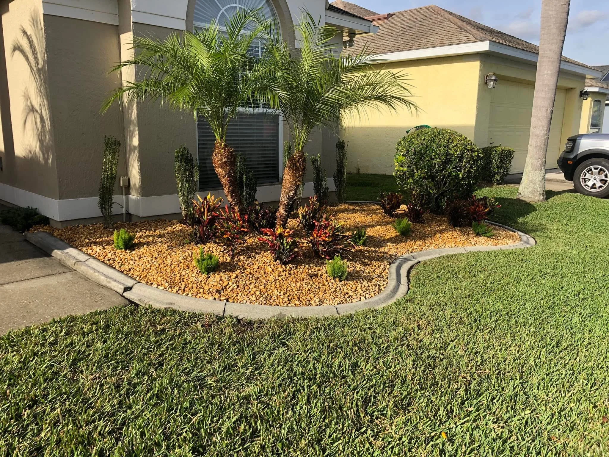 A landscaped front yard with two small palm trees, various bushes, and colorful plants surrounded by yellow gravel, with a concrete border and green lawn, next to a beige house and a driveway with a vehicle.
