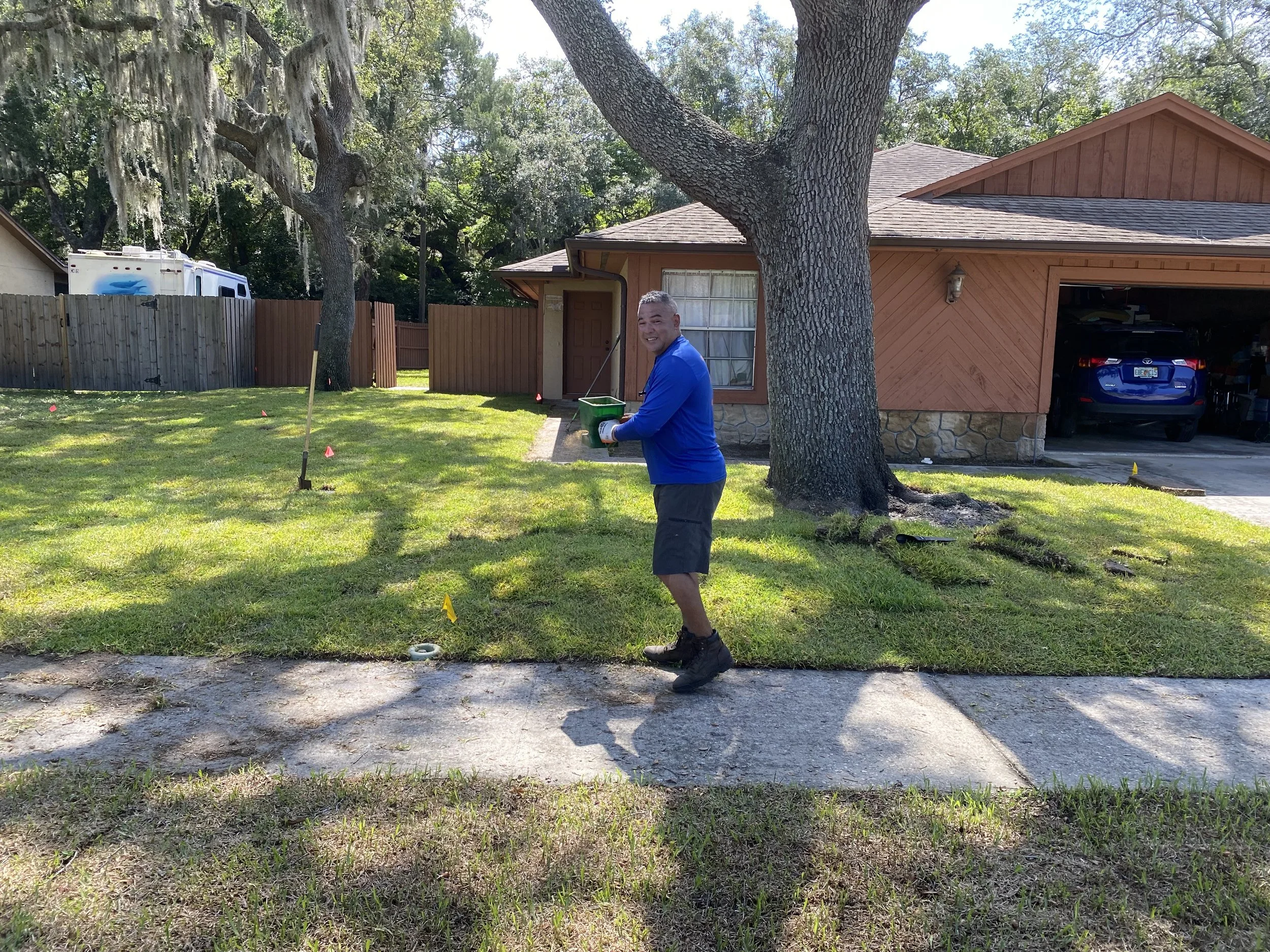 A man standing on a sidewalk holds a green bucket, smiling, in front of a house with a large tree. The yard has freshly cut grass with small markers around it, and the house has a closed garage with a blue car inside.