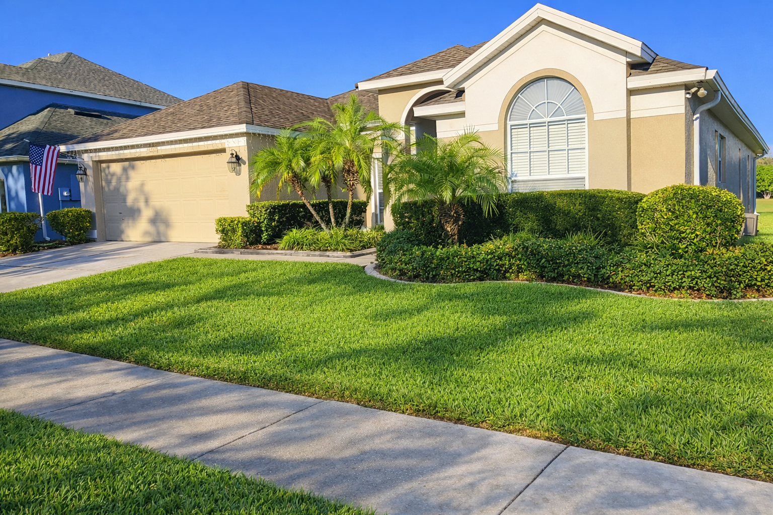 Front view of a single-story house with a well-maintained lawn, palm trees, bushes, and a driveway leading to a garage. The house has beige siding, a large arched window, and an American flag display.