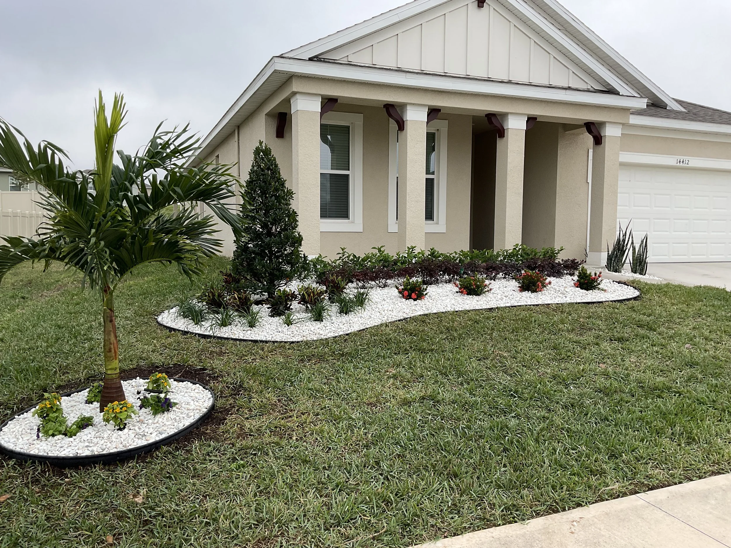 Front yard landscaping of a beige house with a covered porch, featuring white gravel beds with plants, small bushes with red flowers, and a small palm tree.
