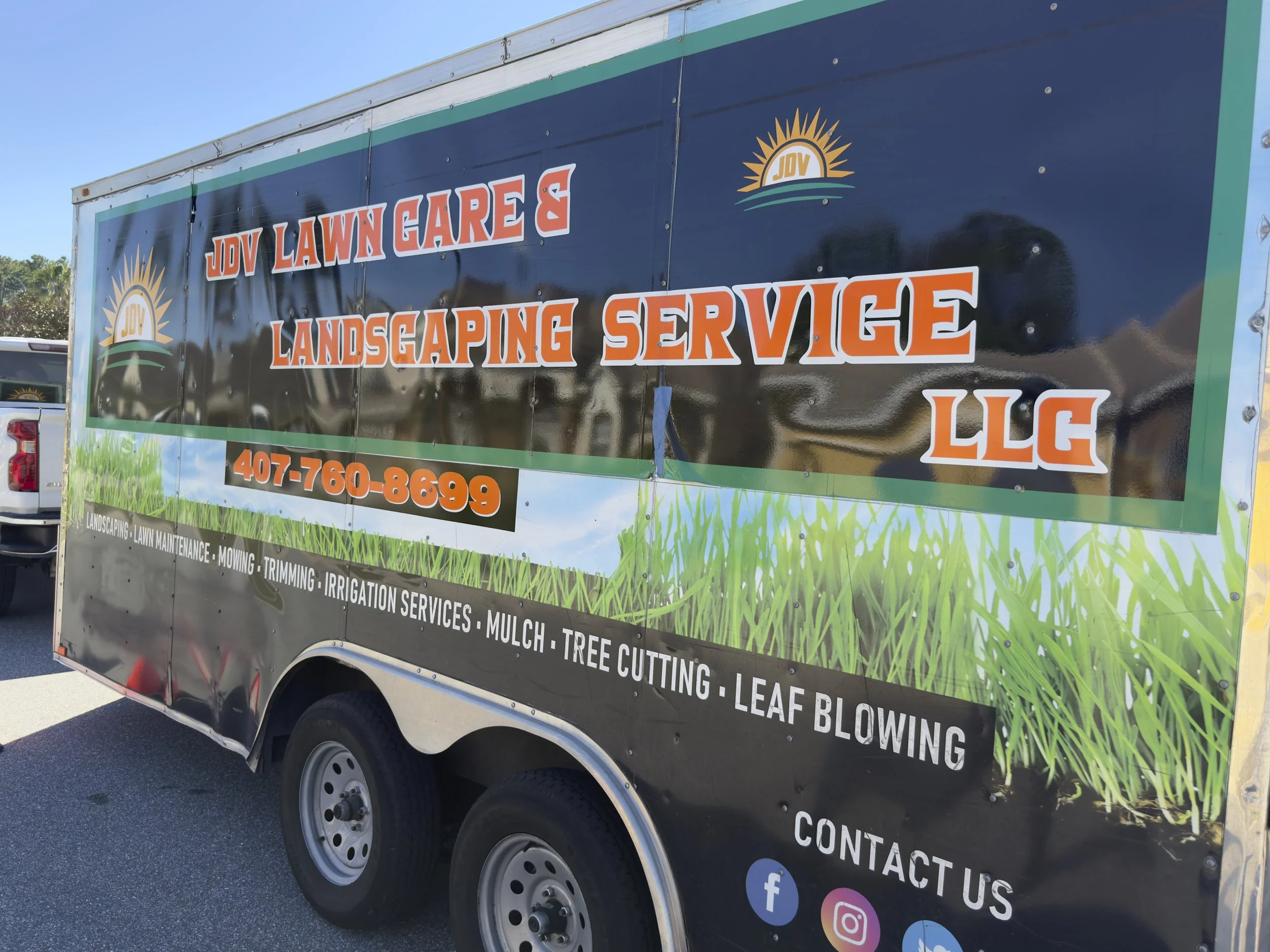 Trailer advertising JDV Lawn Care and Landscaping Service LLC with a black background, orange and white text, and contact information. The trailer also lists services such as lawn maintenance, mowing, trimming, irrigation services, mulch, tree cutting, and leaf blowing, with a grass field and sky in the background.