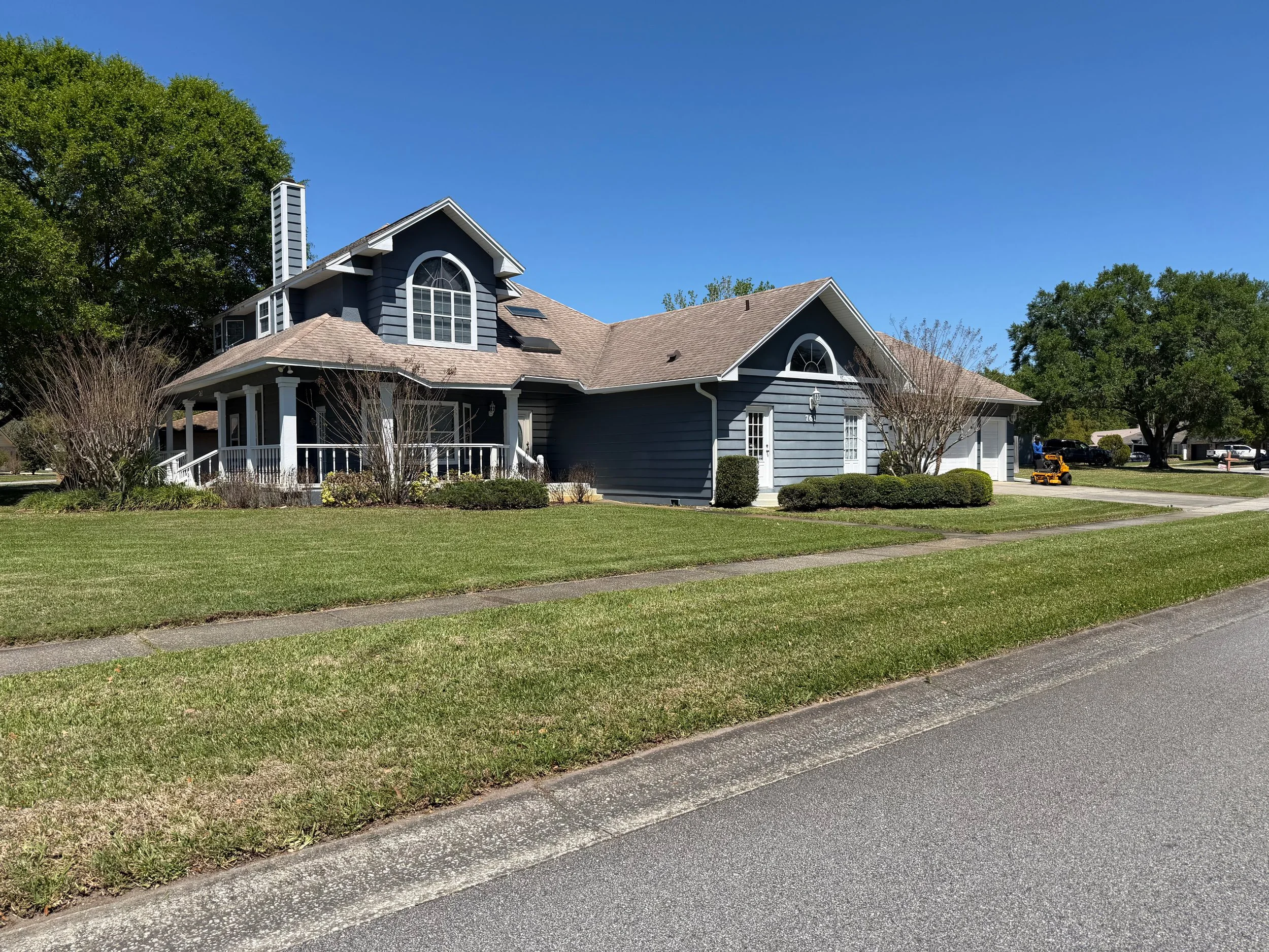 A two-story house painted in blue with white trim in a suburban neighborhood. The house has a pitched roof with dormer windows, a front porch with columns, and a well-maintained lawn with bushes and trees.